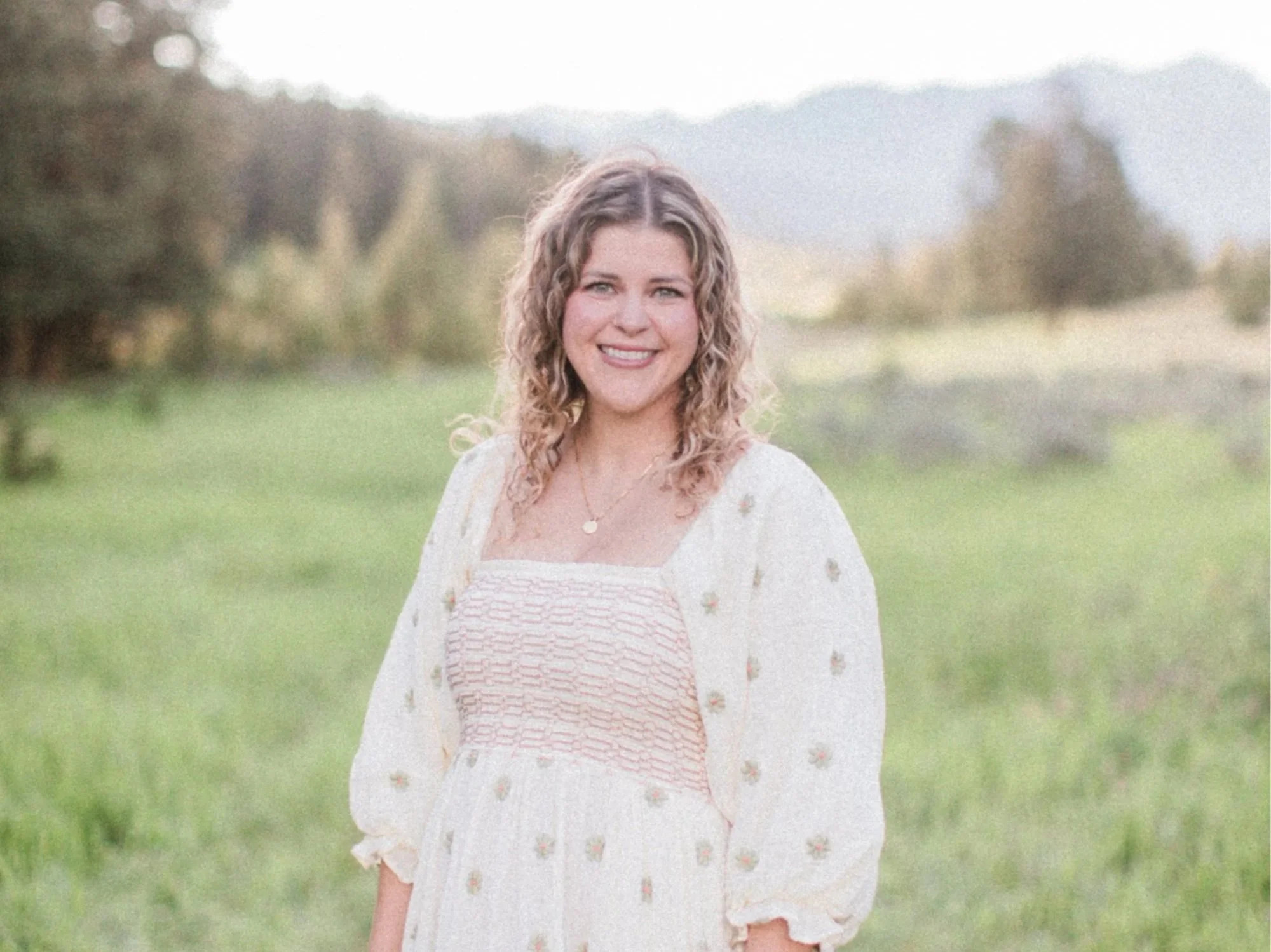 A woman with curly blonde hair smiling outdoors in a green field with trees and mountains in the background.