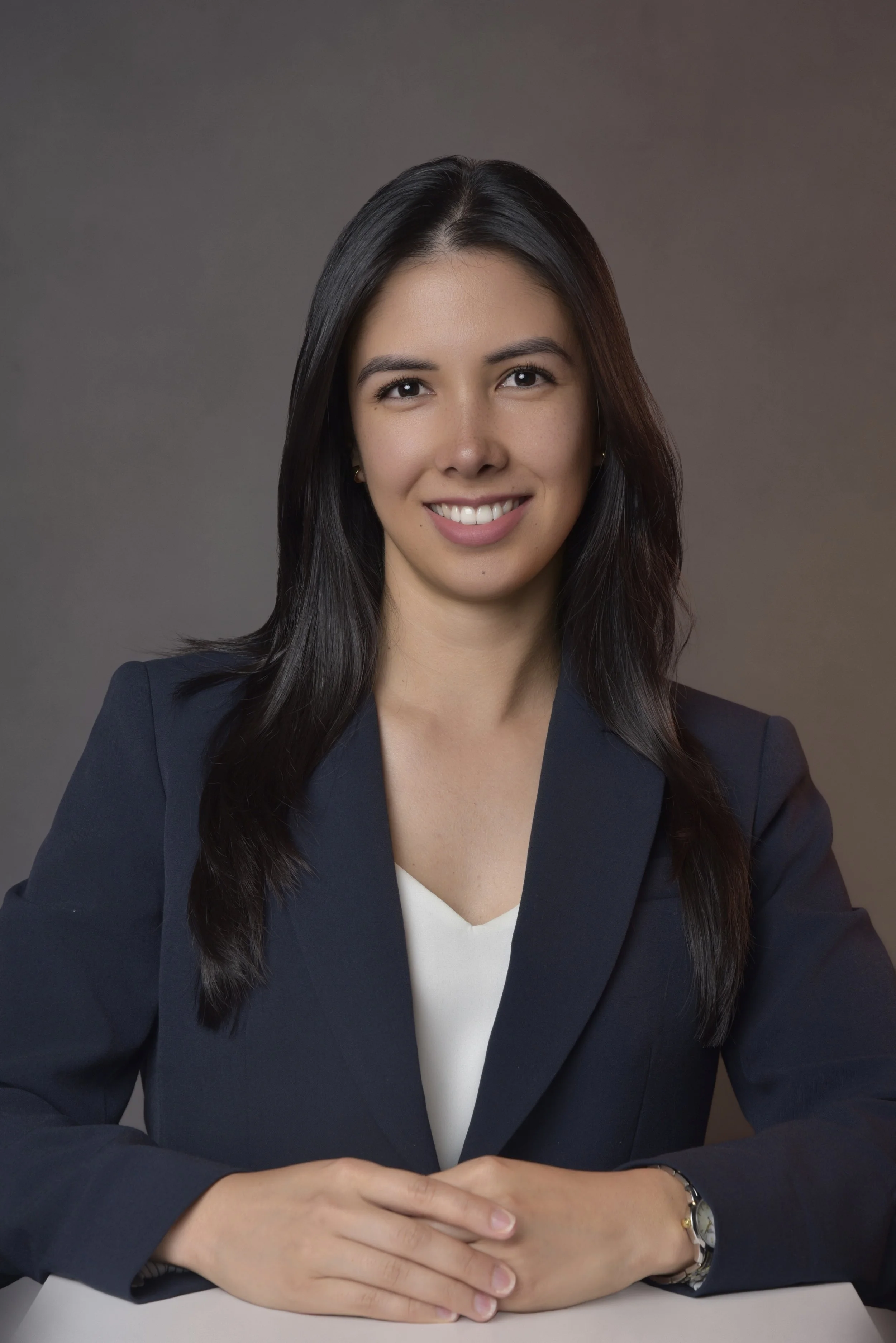Professional portrait of a young woman with long dark hair smiling, wearing a dark blazer and white top, seated at a desk against a neutral background.