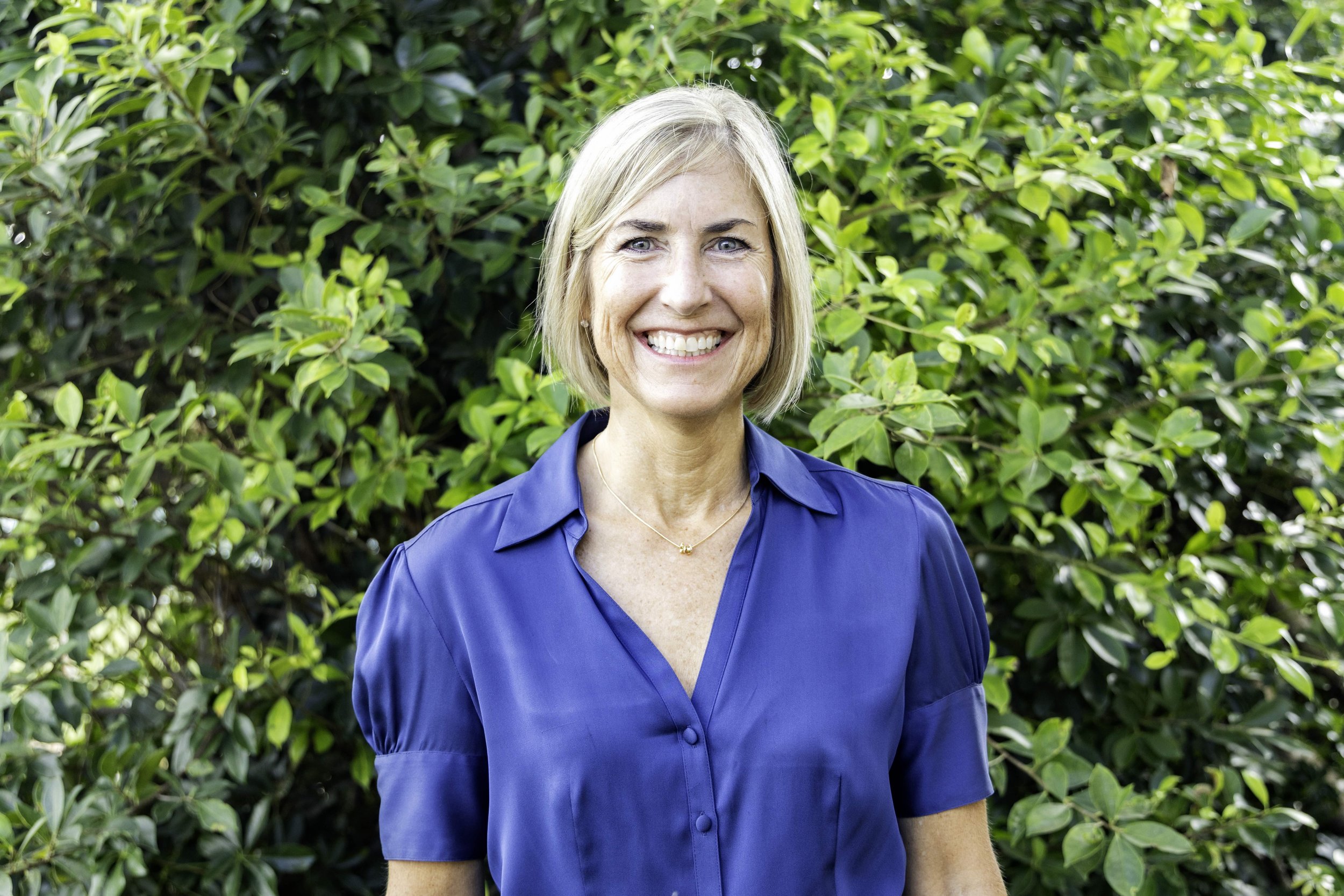Smiling woman with shoulder-length blonde hair wearing a blue shirt, standing in front of green leafy bushes.