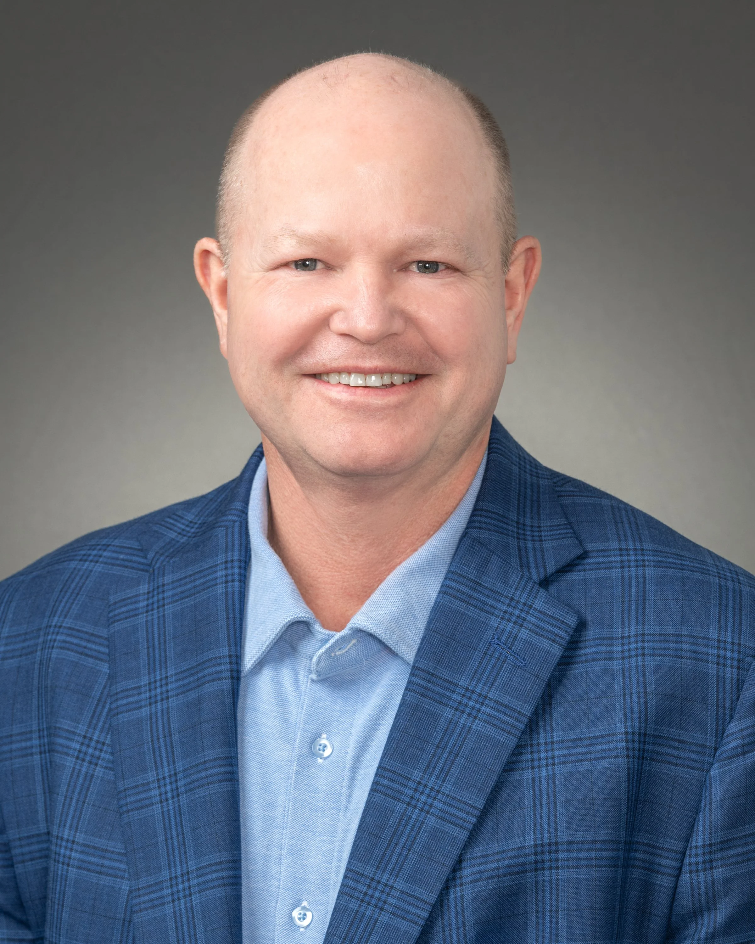A headshot of a smiling bald man wearing a blue checked blazer and a light blue shirt against a gray background.