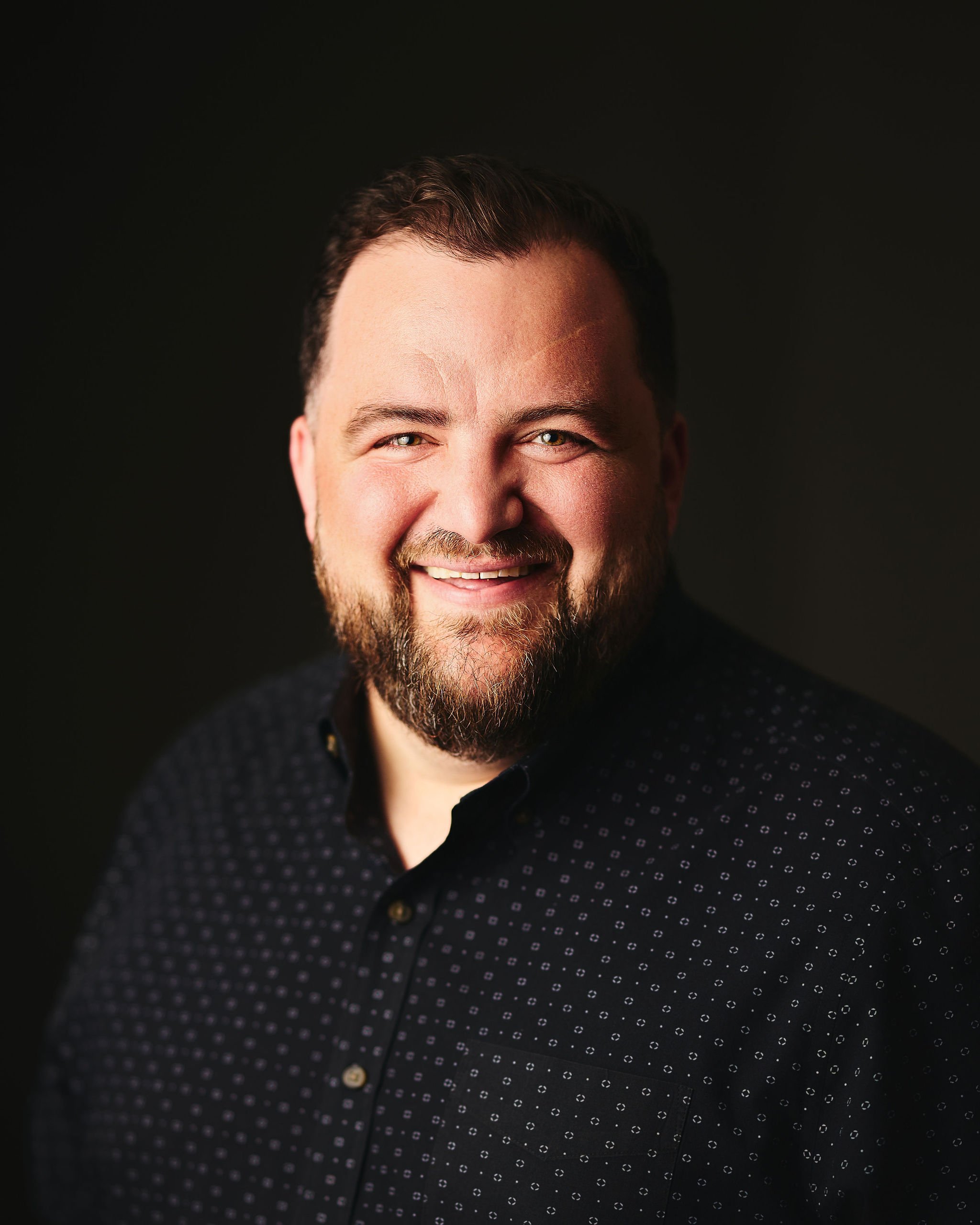 A smiling man with a beard and mustache, wearing a black button-up shirt with small white patterns, against a dark background.