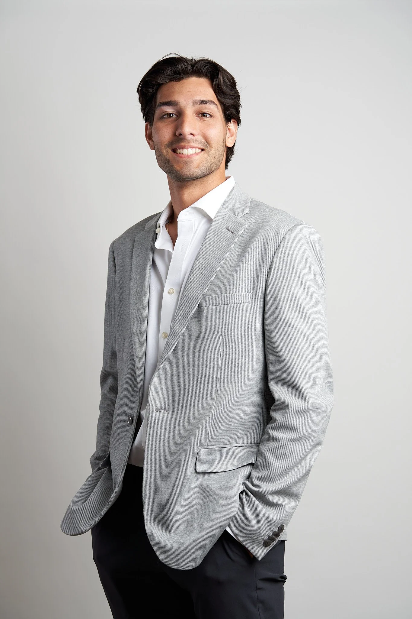 A smiling young man with dark, wavy hair, wearing a light gray suit jacket over a white dress shirt, with his right hand in his pocket, standing against a plain light gray background.