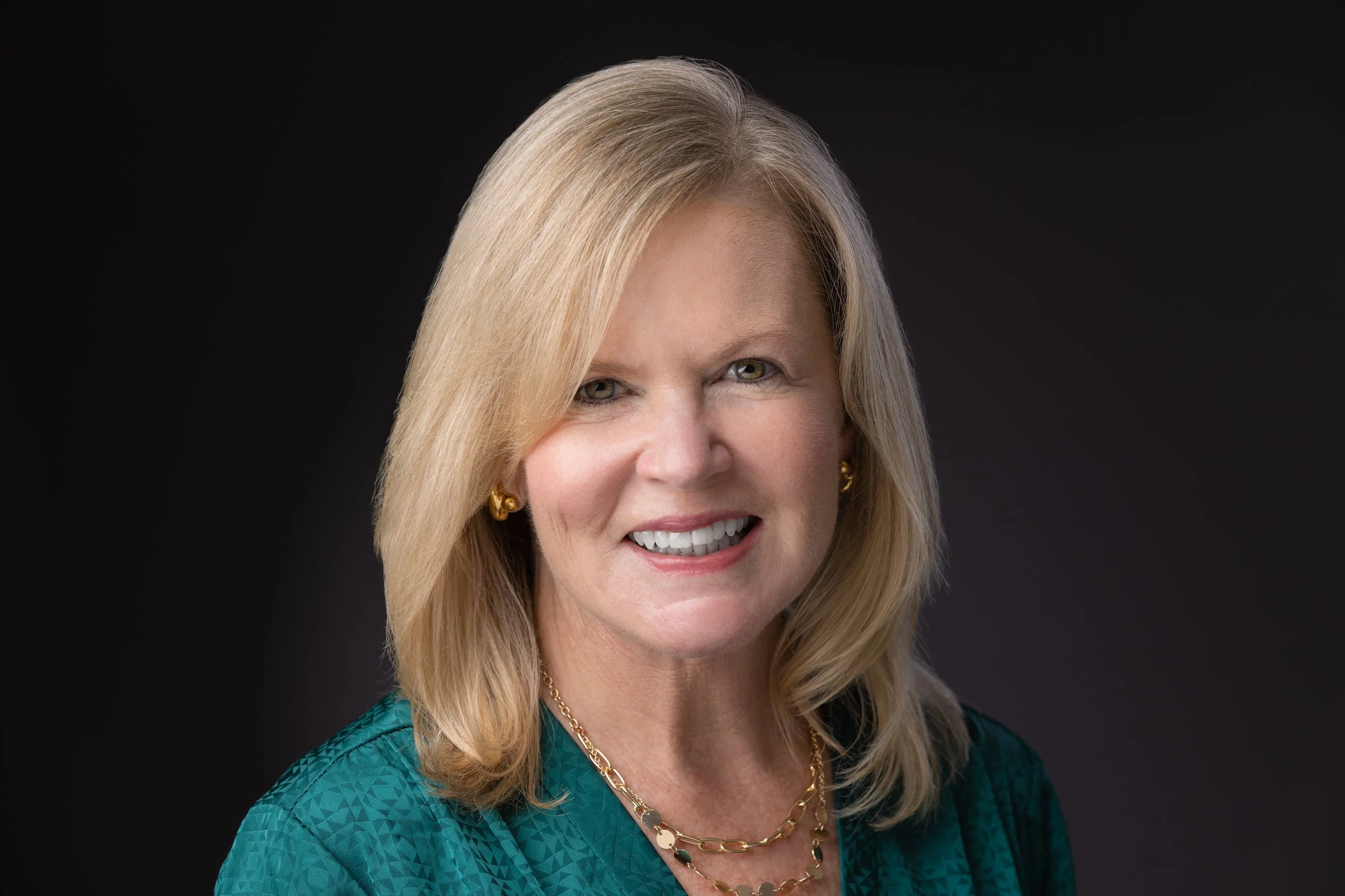 A portrait of a smiling middle-aged woman with blonde hair, wearing a teal blouse and gold jewelry, against a dark background.