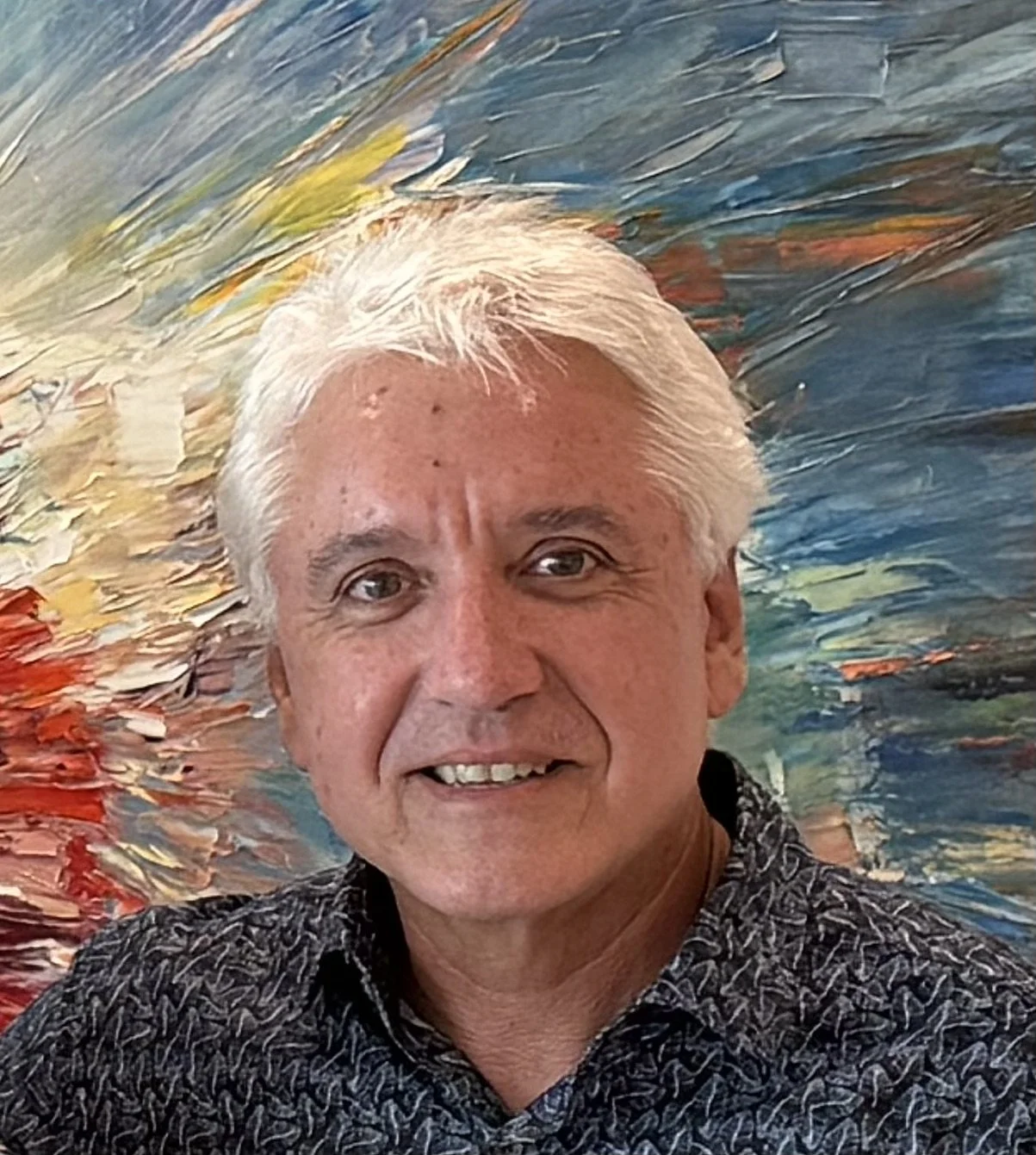 A smiling older man with white hair standing in front of a colorful, abstract painting with swirling red, yellow, blue, and green strokes.