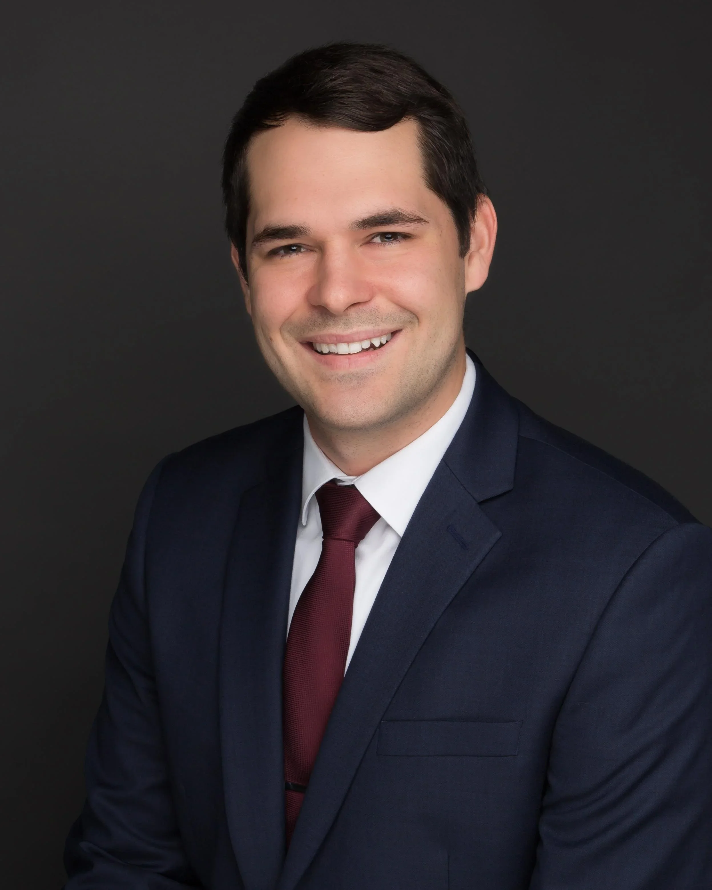 Professional headshot of a man wearing a navy suit, white shirt, and burgundy tie, smiling against a dark background.