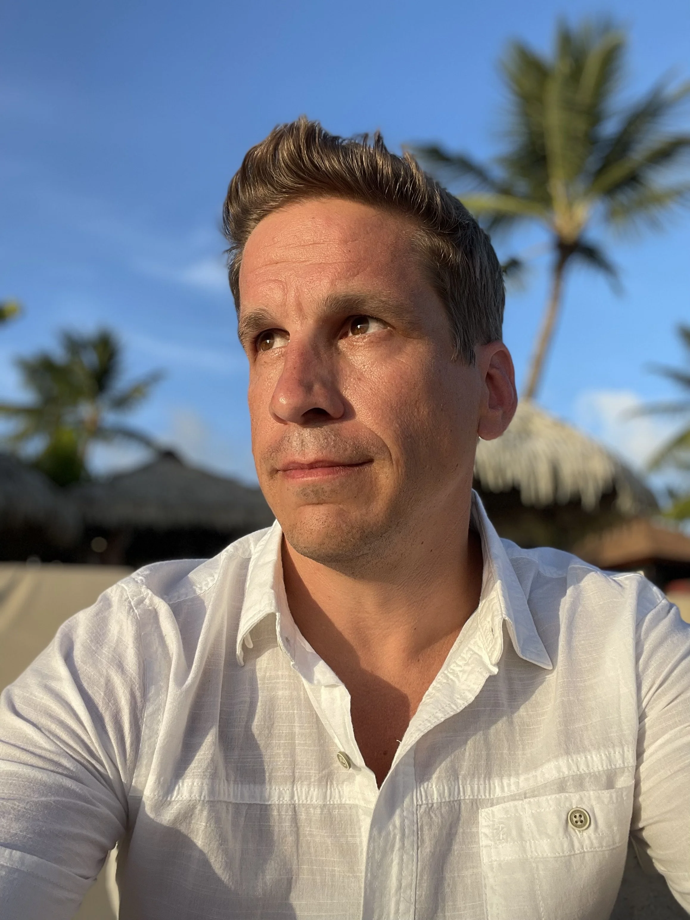 A man in a white shirt is outdoors with a beach background, palm trees, and thatched huts during sunset.