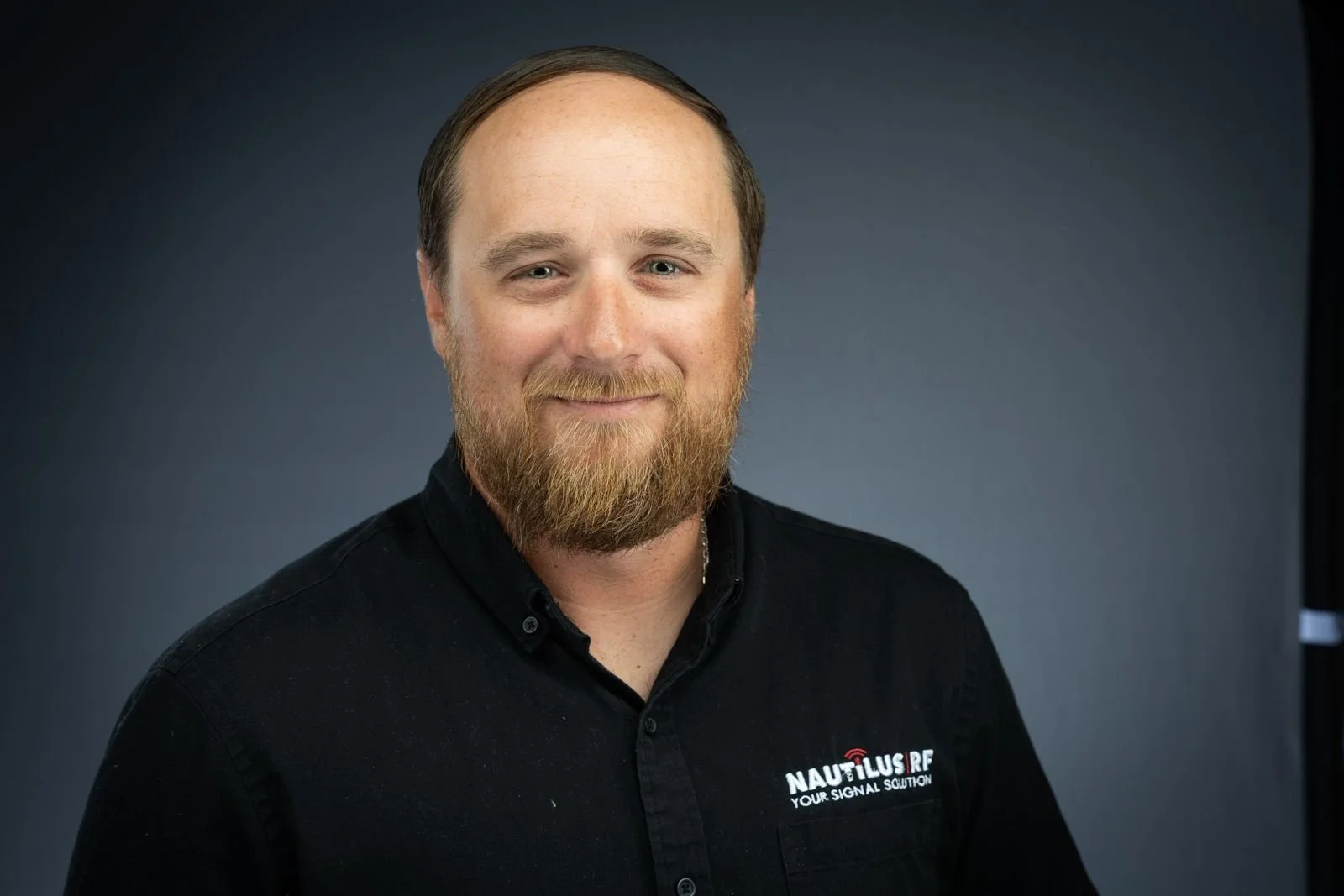 A man with a beard and short hair wearing a black button-up shirt with the logo 'NAUTILUS RF YOUR SIGNAL SOLUTION' on the chest, smiling against a dark gradient background.