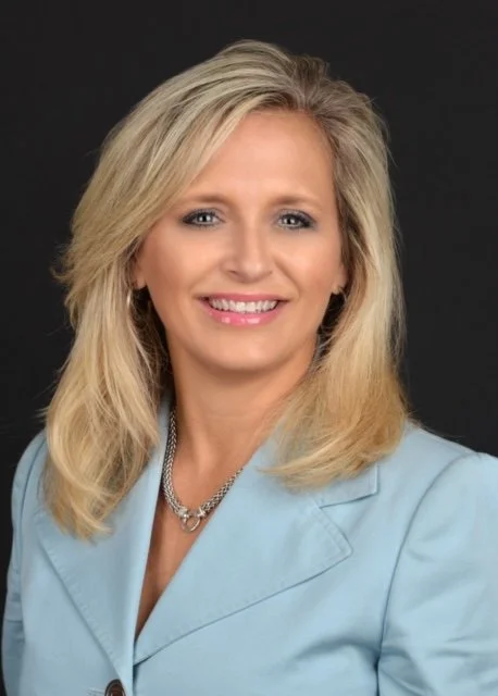 Professional headshot of a smiling blonde woman in a light blue blazer with a silver necklace against a black background.