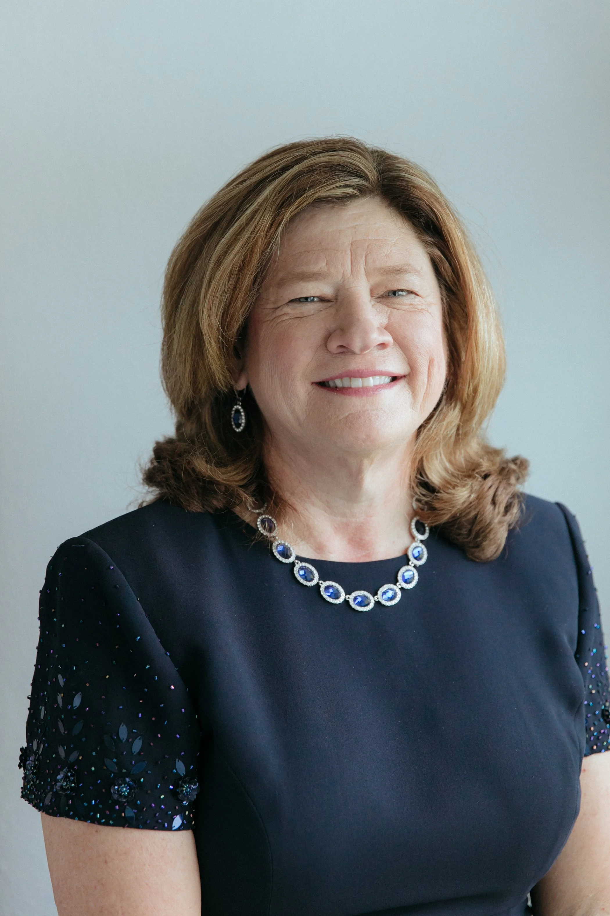 A woman with shoulder-length light brown hair wearing a navy blue dress with embroidered sleeves, a matching blue gemstone necklace, and earrings, smiling at the camera against a plain light background.