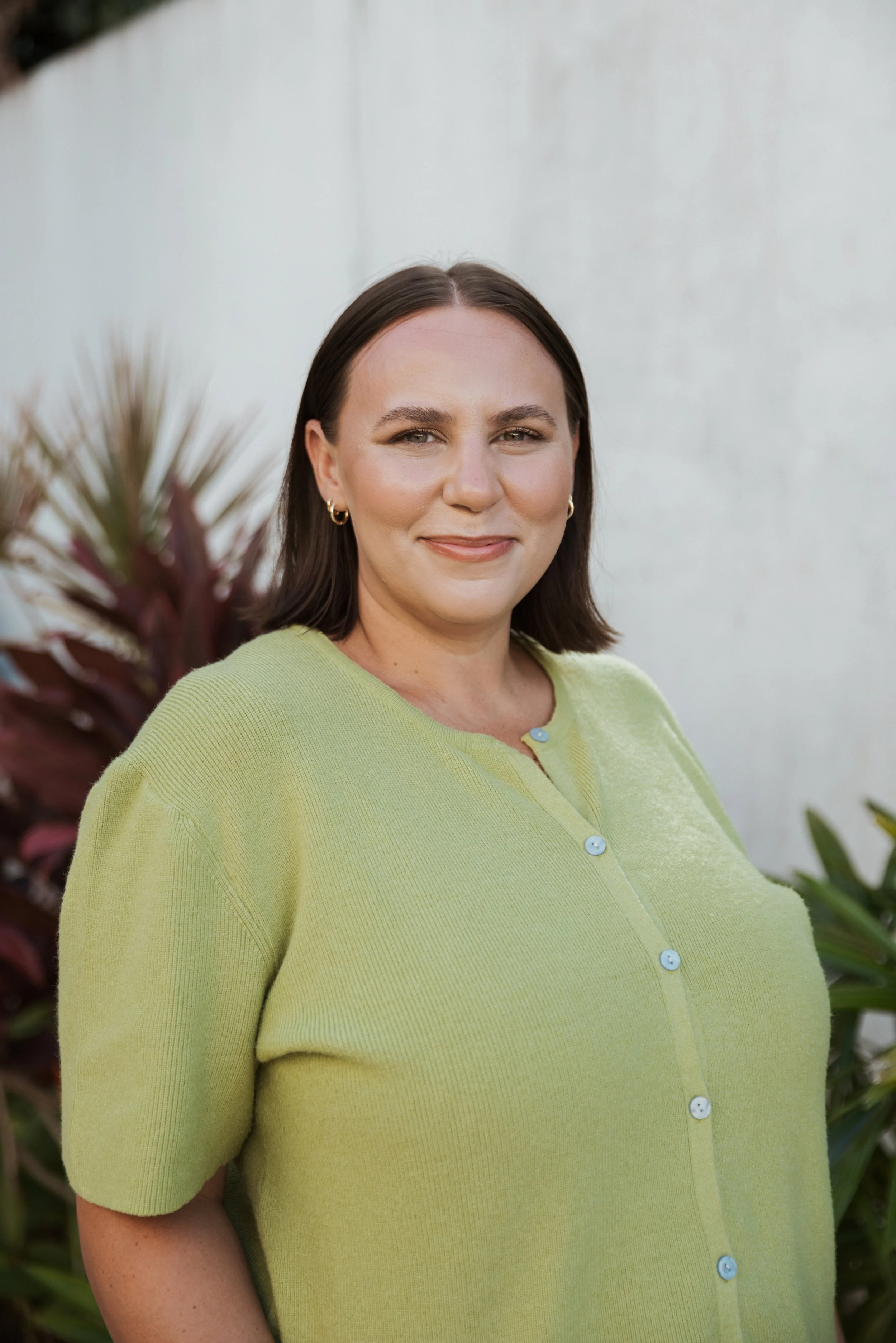 A woman with shoulder-length dark hair, wearing a light green cardigan and gold hoop earrings, standing outdoors in front of a white wall and tropical plants.