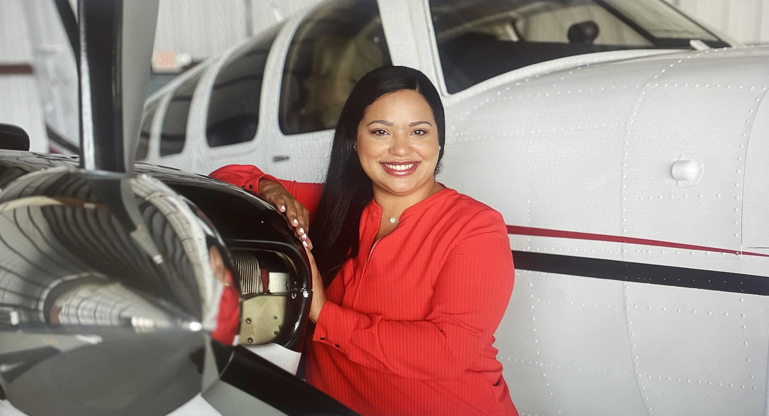 A woman in a red shirt leaning on a small aircraft in a hangar, smiling at the camera.
