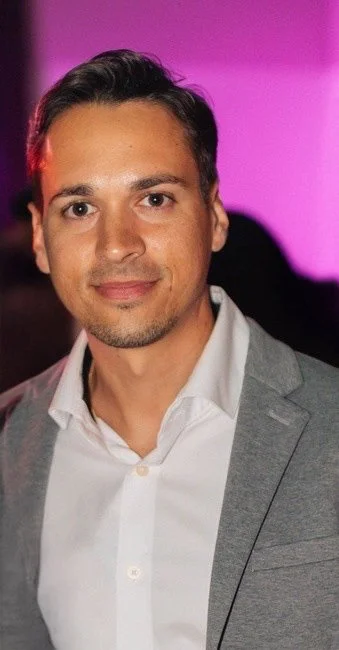 A close-up of a man in a gray suit and white dress shirt, with a pink and purple blurred background.