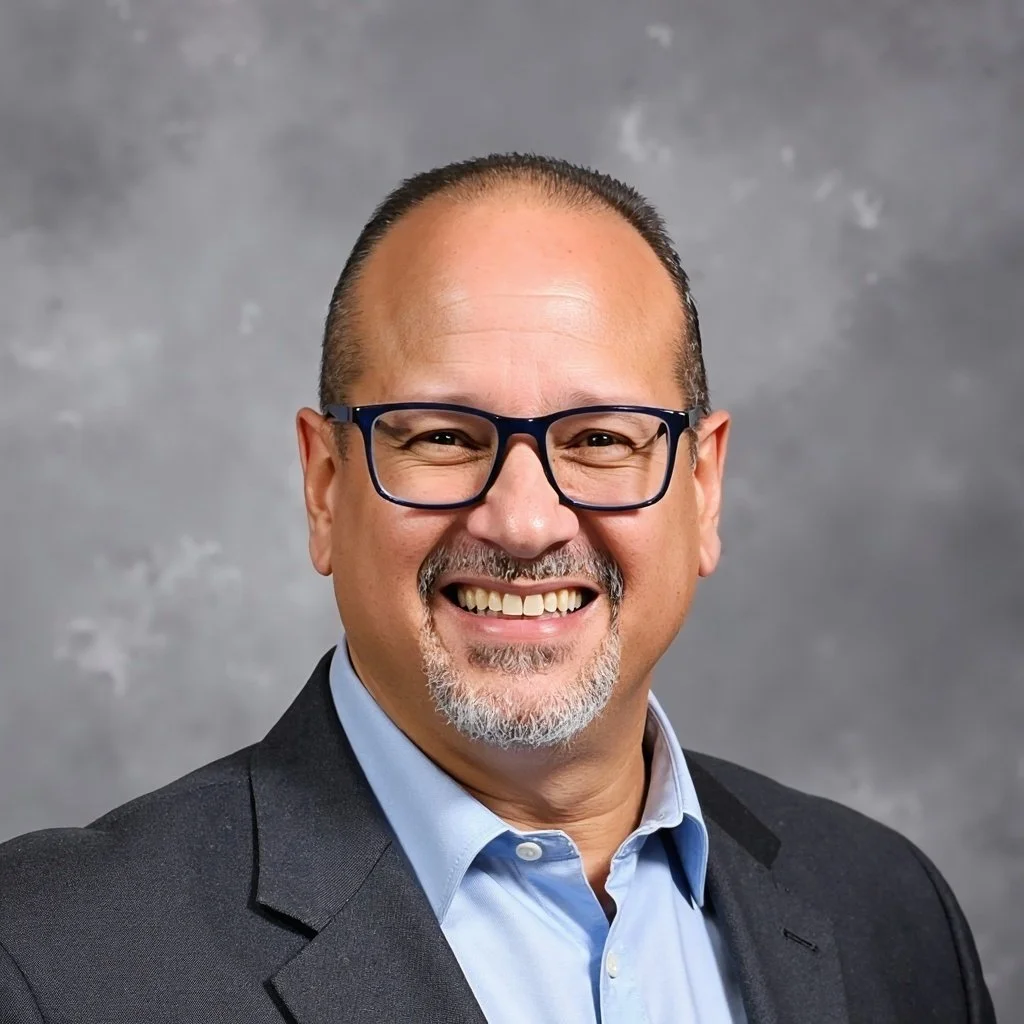 Portrait of a smiling man with glasses, wearing a suit jacket and a light blue dress shirt.