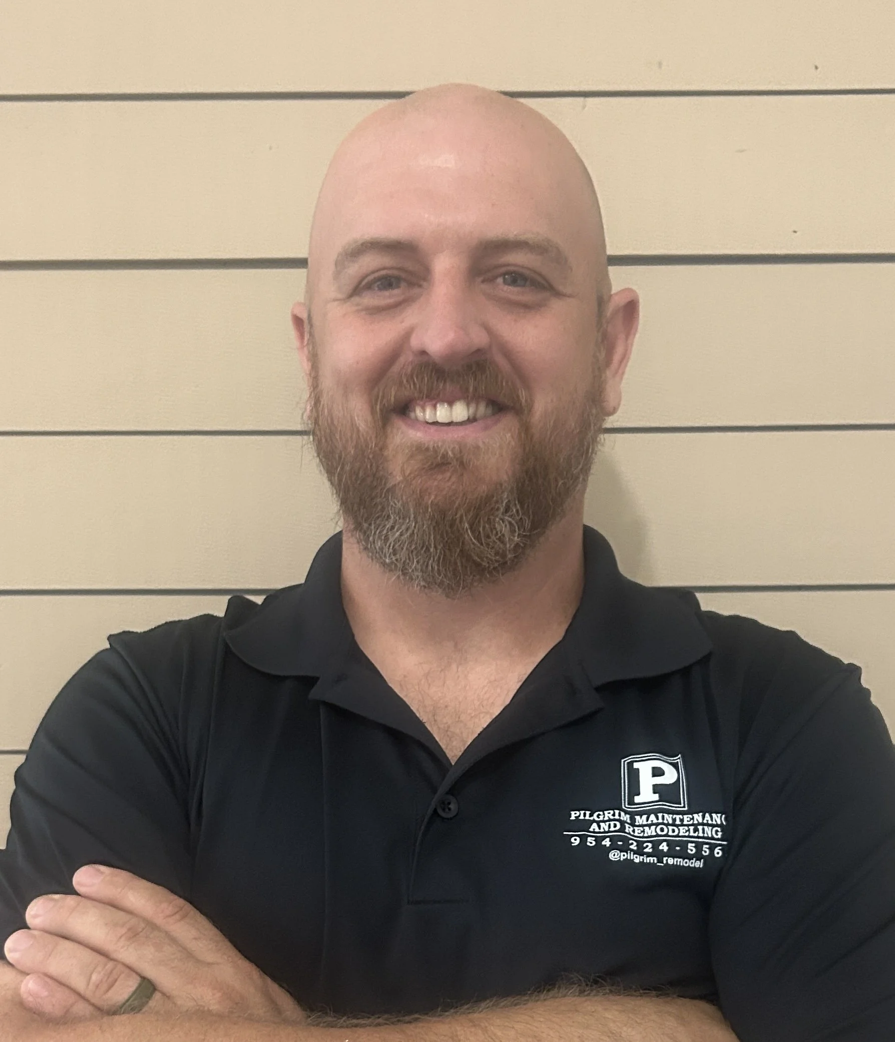 A smiling man with a beard and mustache, wearing a black polo shirt with a company logo, standing against a beige horizontal panel wall.