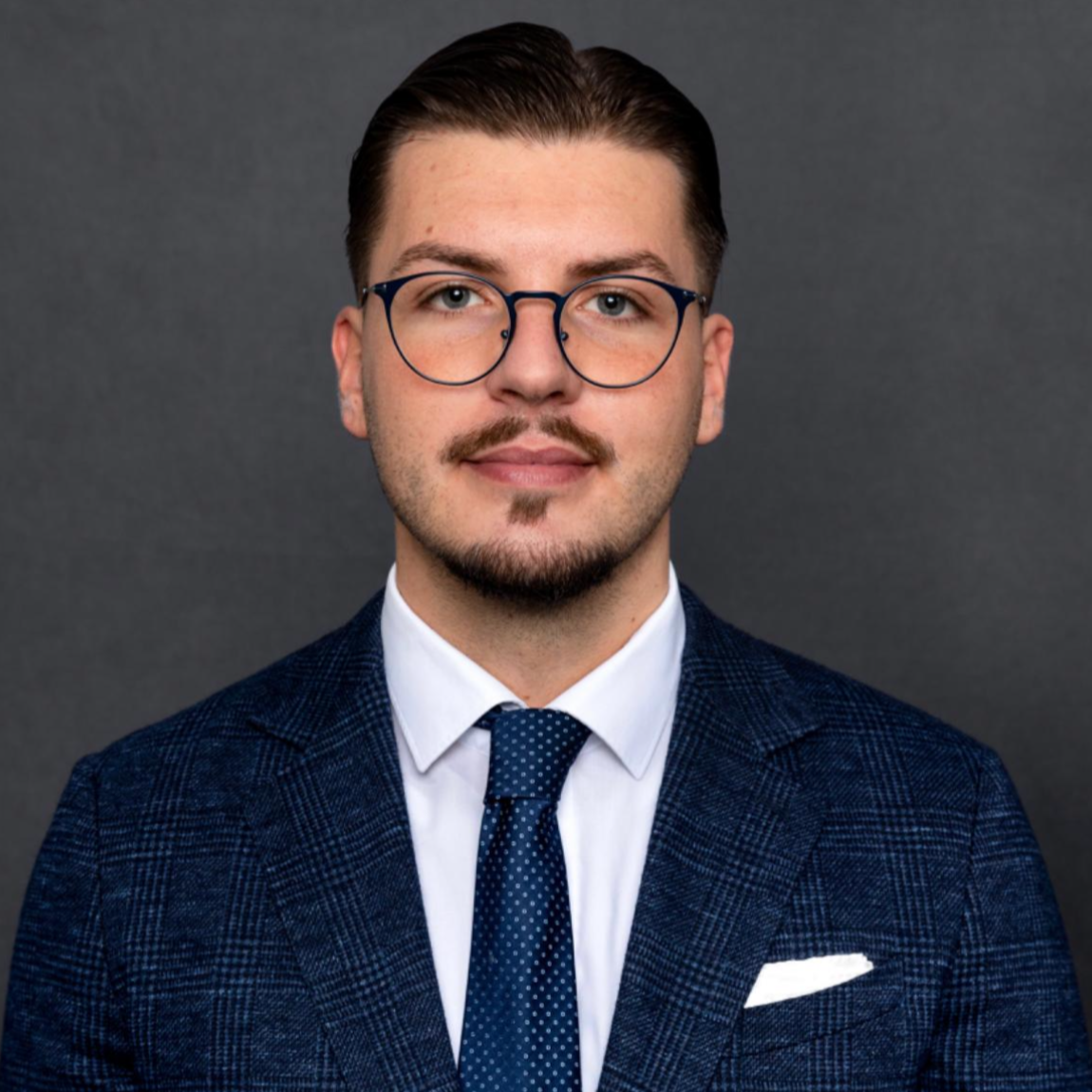 Headshot of a man wearing glasses, a dark patterned blazer, a white shirt, and a dark dotted tie, against a dark grey background.