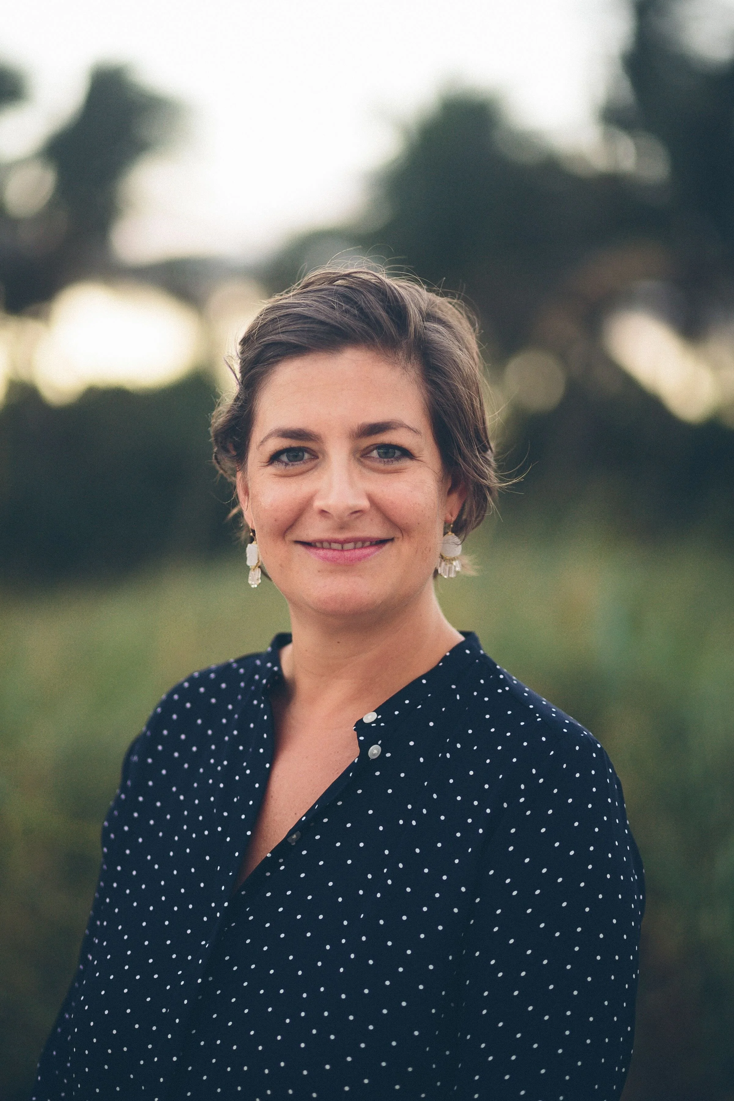 A woman with short brown hair wearing a black blouse with white polka dots and dangling earrings stands outdoors with a blurred background of trees and sky.