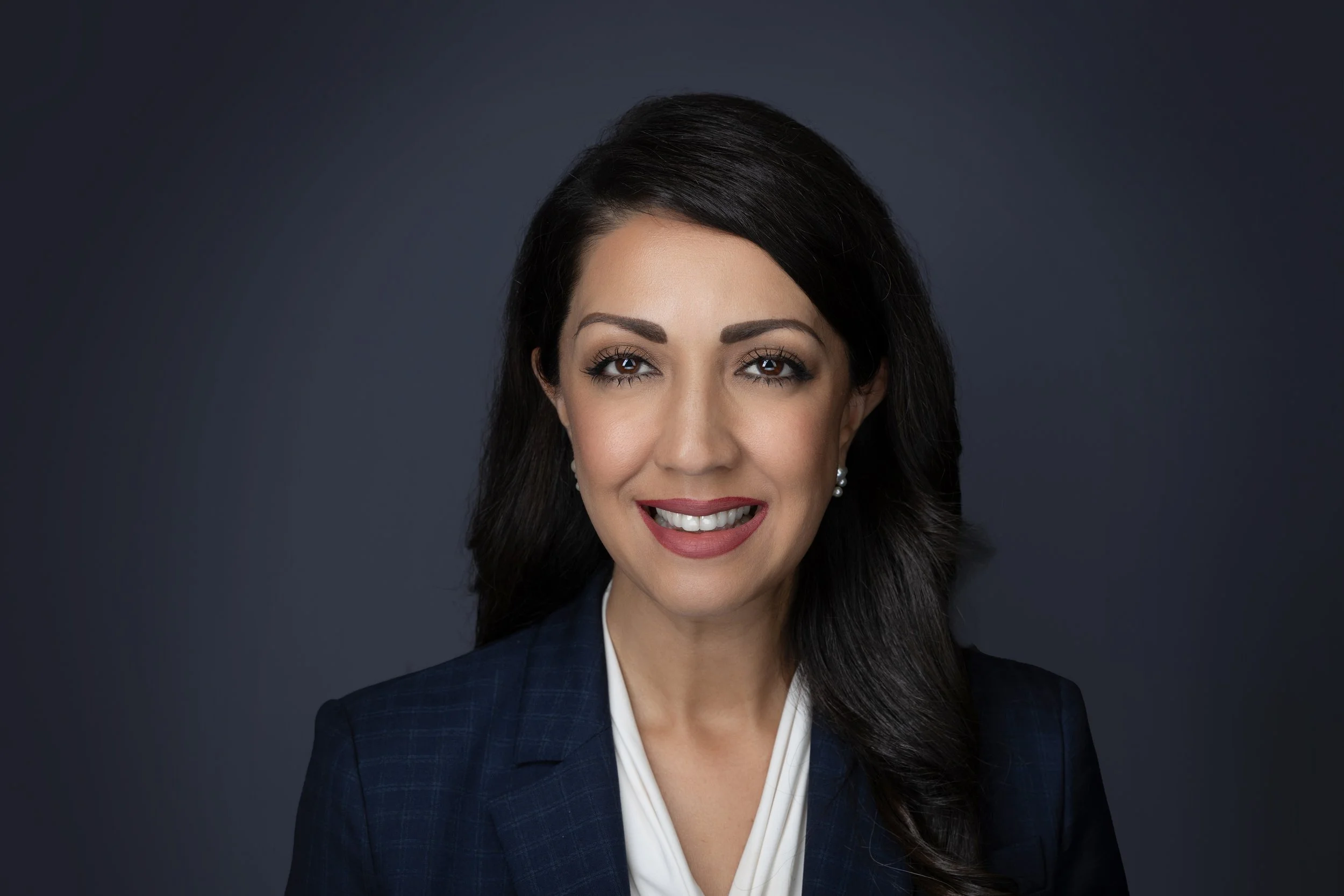 A professional woman with long dark hair, wearing a dark blazer and pearl earrings, smiling against a dark gray background.
