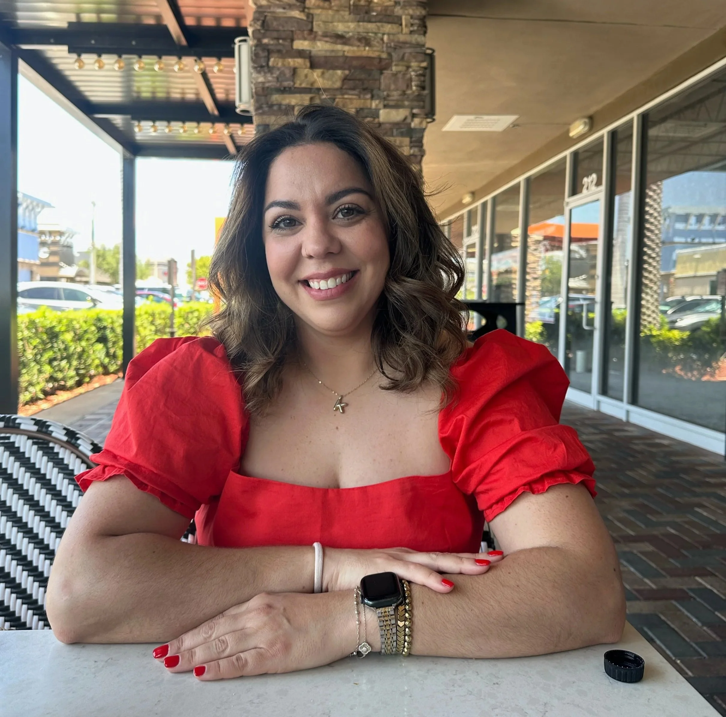 A woman sitting at a table in an outdoor seating area of a restaurant or cafe, smiling at the camera with her arms crossed on the table, wearing a red puff-sleeve top, jewelry, and a smartwatch.