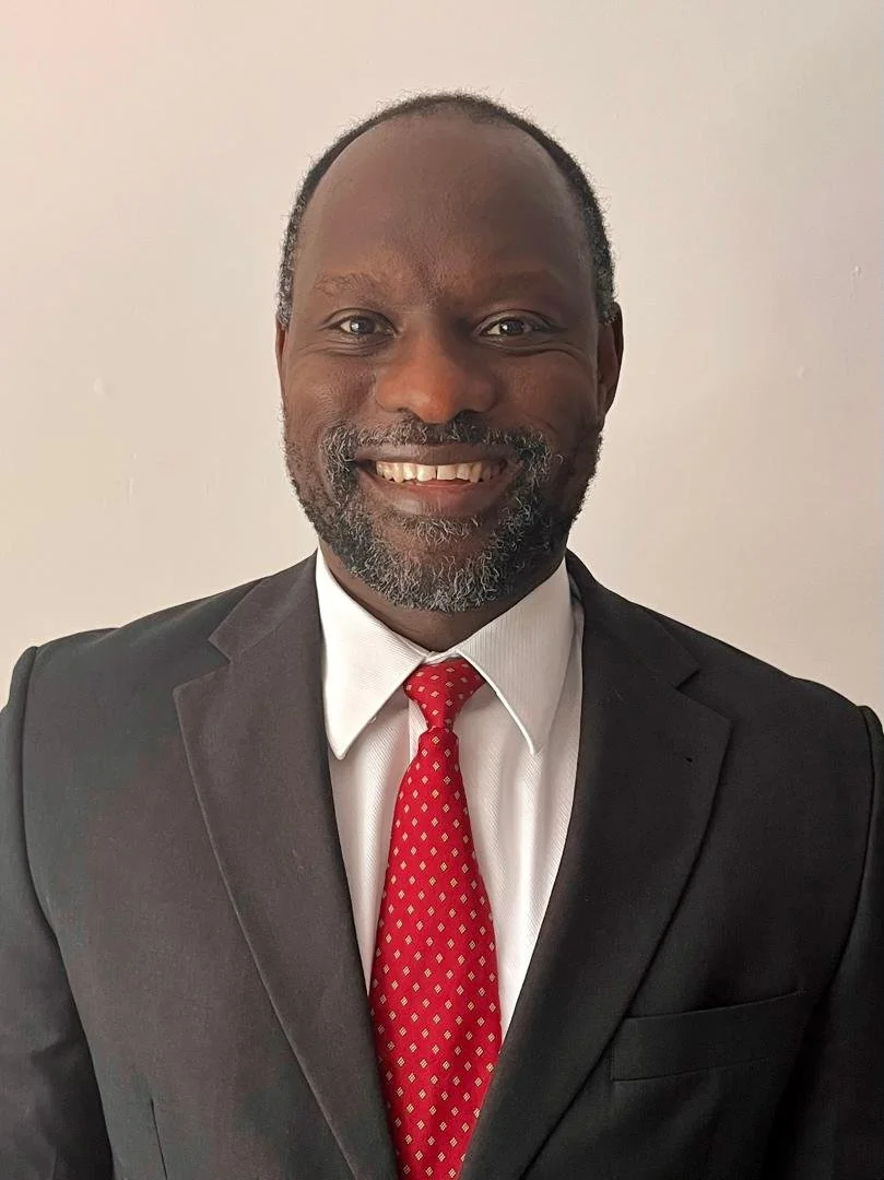 A smiling man wearing a black suit, white dress shirt, and red patterned tie, standing against a plain light-colored background.