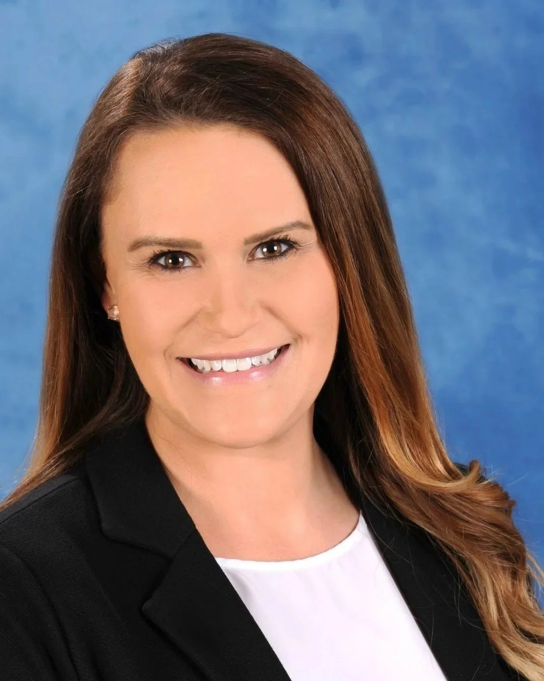 Professional headshot of a woman with long, wavy brown hair, smiling, wearing a black blazer and white top, against a blue background.