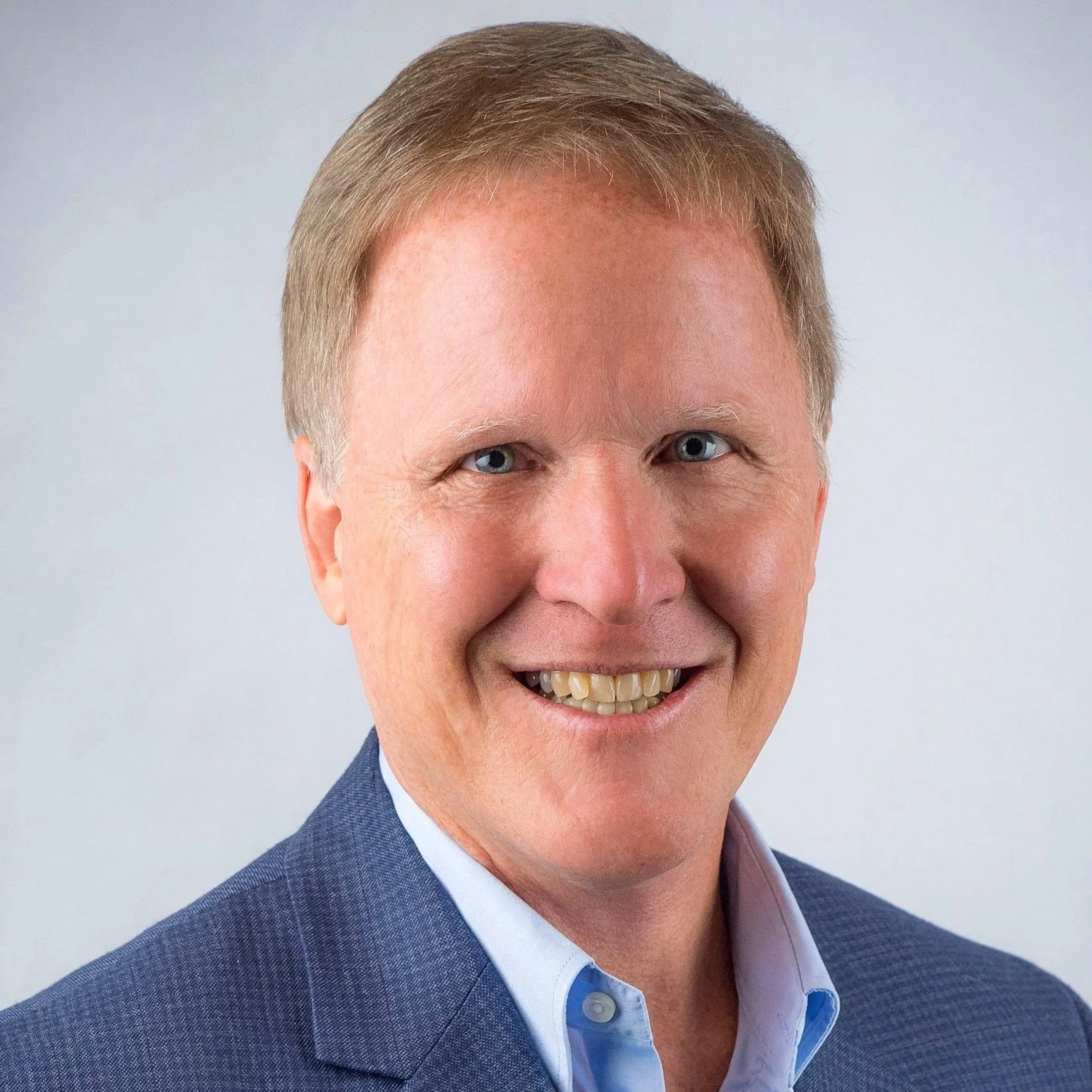 Headshot of a smiling man with blond hair, blue eyes, wearing a blue suit and light blue shirt, against a plain grey background.