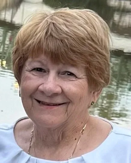 Close-up of an elderly woman with short, reddish hair, smiling outdoors near a body of water.
