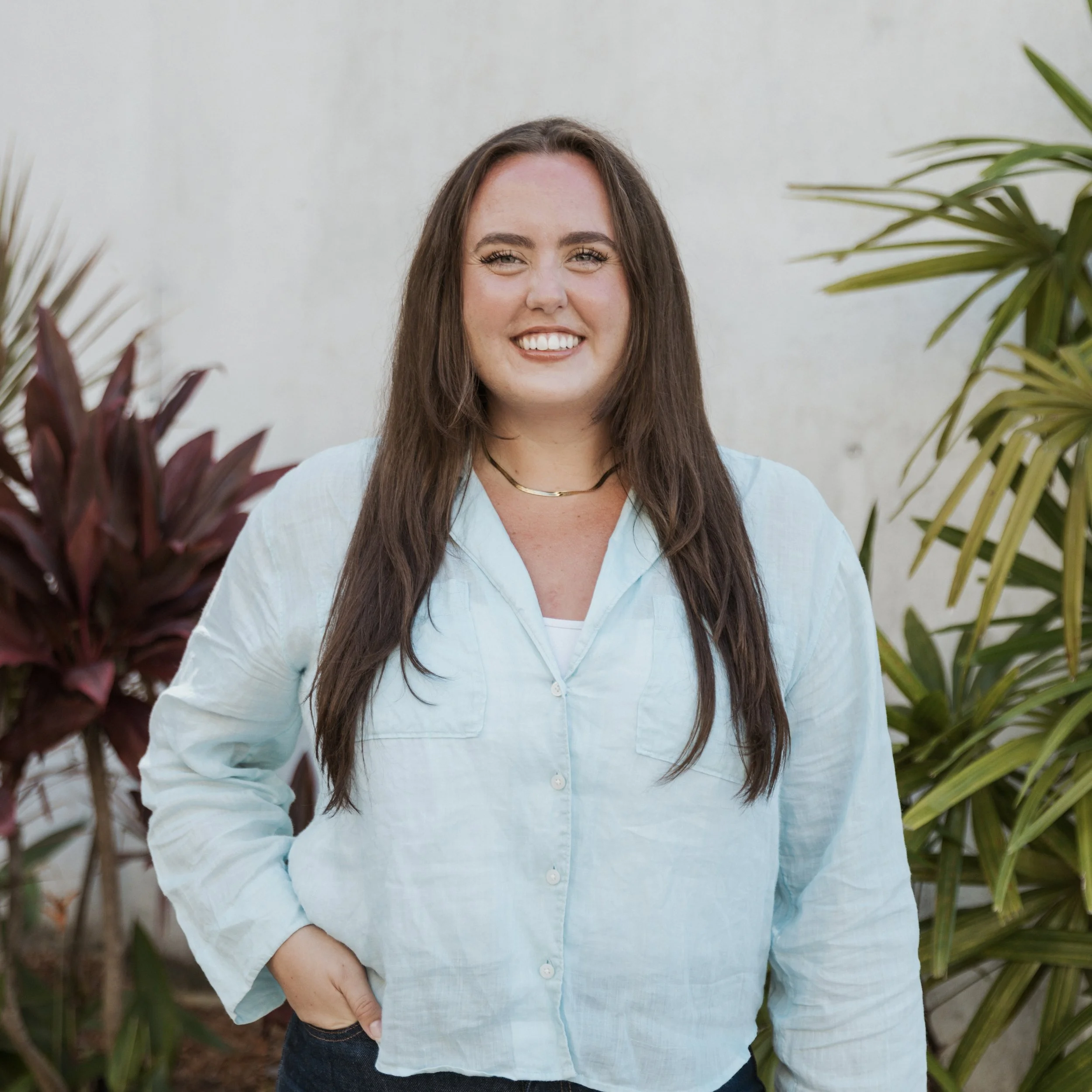 A young woman with long brown hair smiling in front of green and maroon plants on a light-colored wall.