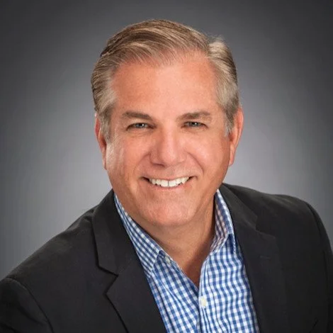Portrait of a smiling middle-aged man with gray hair, wearing a dark blazer over a blue checkered shirt, against a neutral gray background.