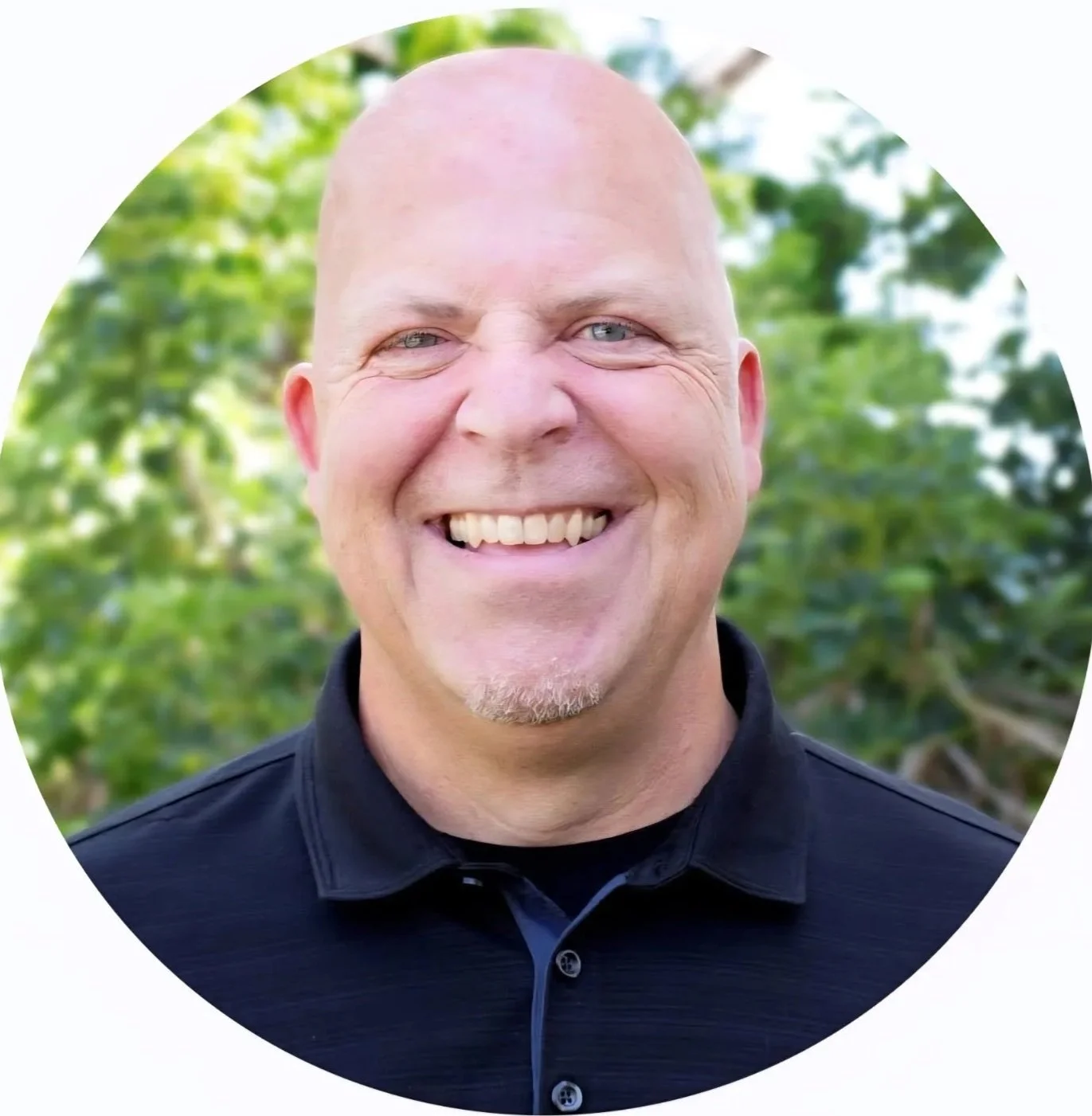 A smiling bald man with a goatee and blue eyes, wearing a black collared shirt, outdoors with green foliage in the background.