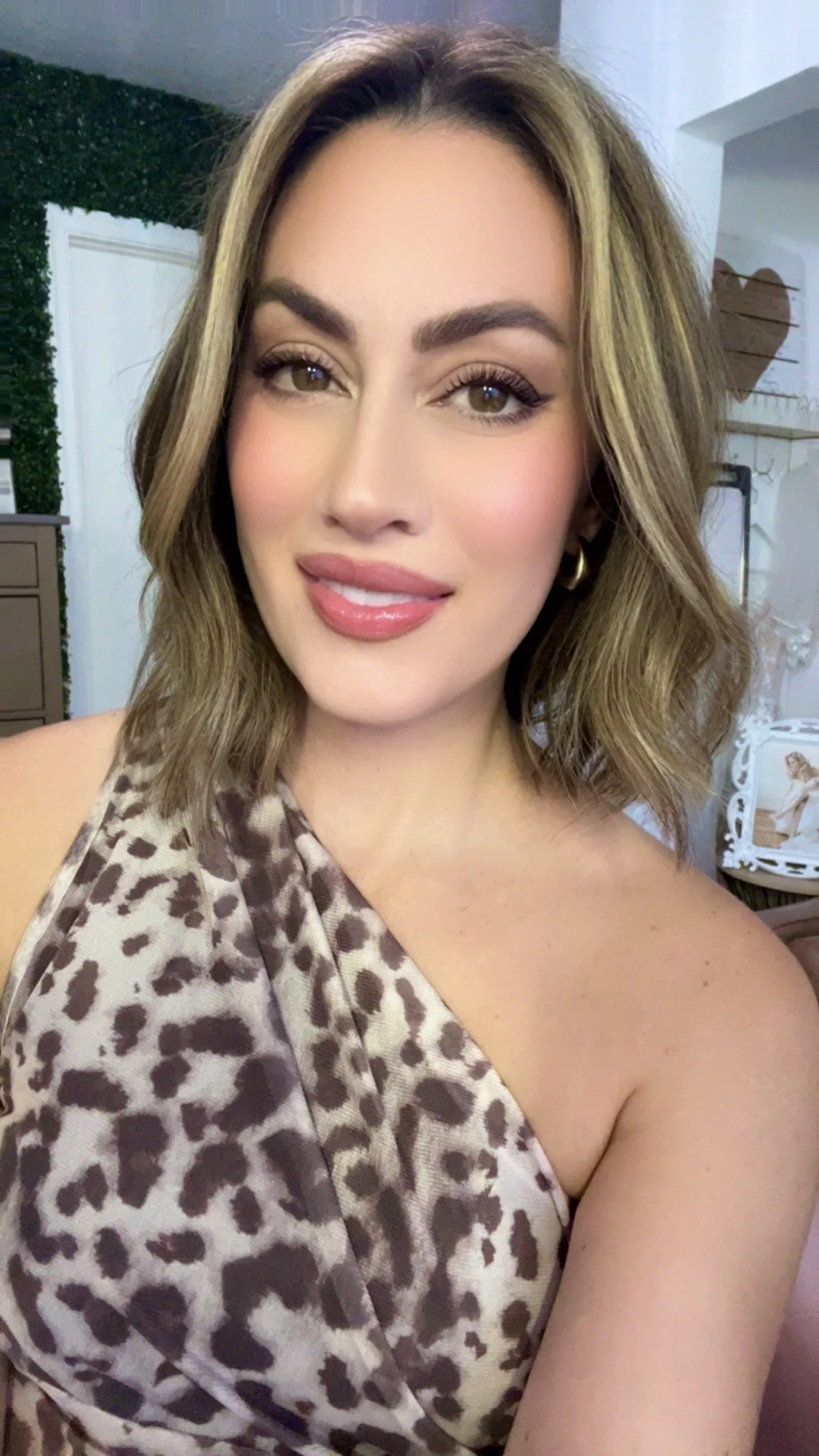 Close-up of a young woman with shoulder-length wavy light brown hair, wearing a leopard print top, smiling with pink lipstick, gold hoop earrings, and natural makeup, indoors with decorative elements in the background.