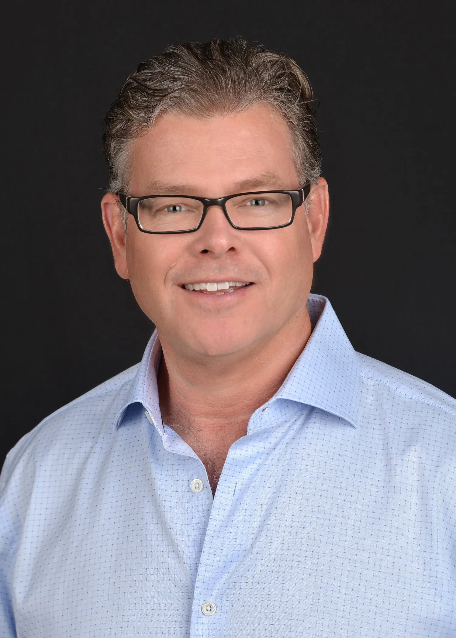 Headshot of a middle-aged man wearing glasses and a light blue collared shirt, smiling, against a black background.