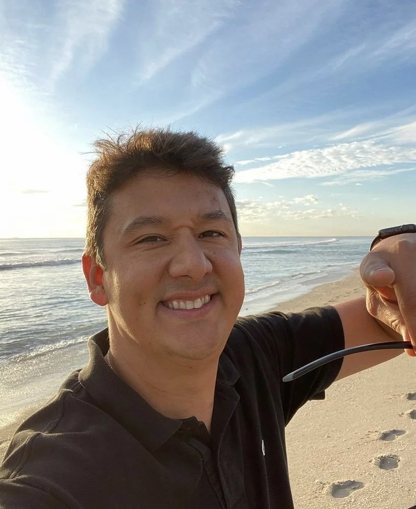A man smiling and taking a selfie on a beach, with the ocean and a partly cloudy sky in the background.