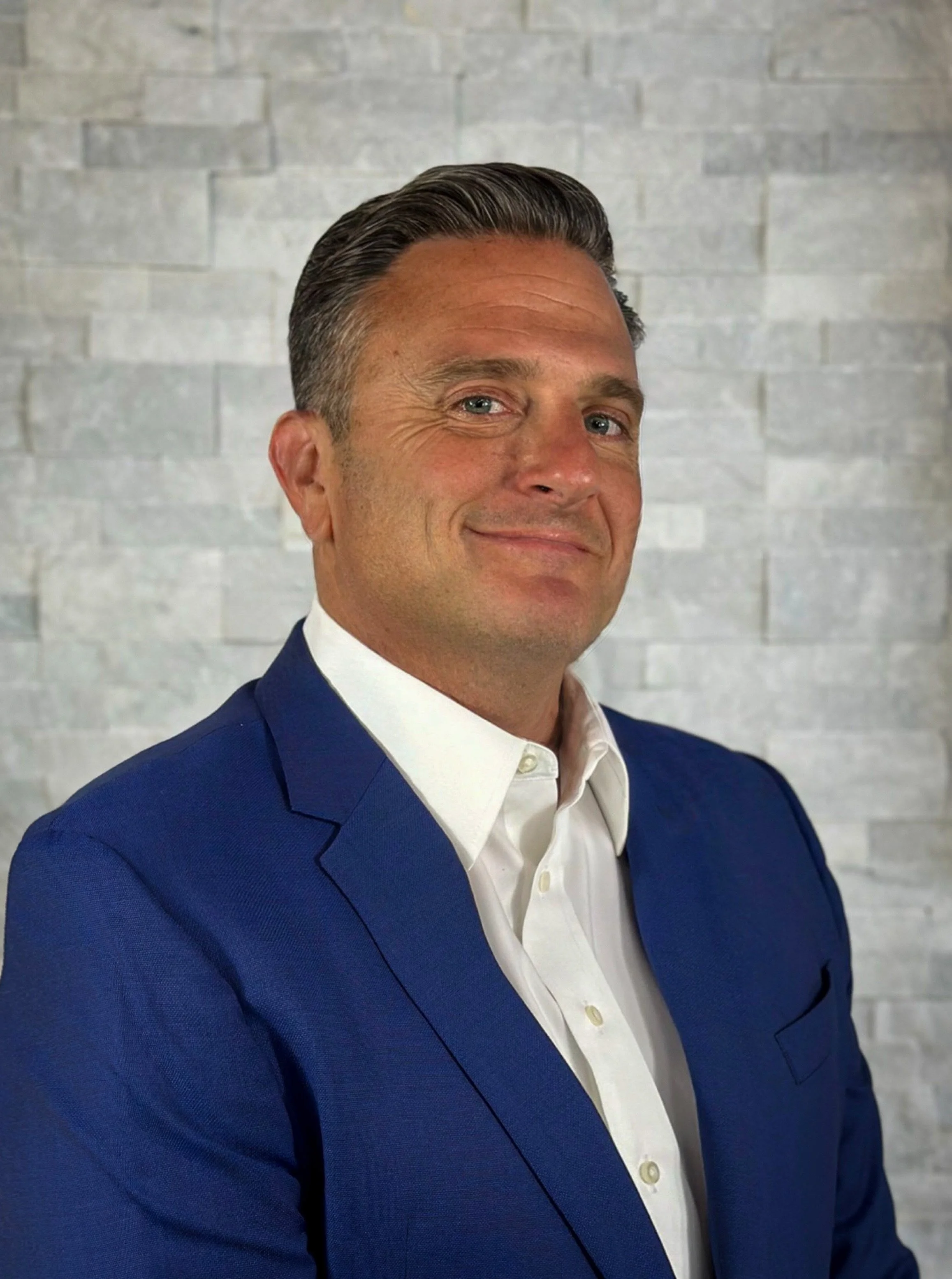 A man wearing a blue suit and white shirt, smiling, standing in front of a light gray brick wall.