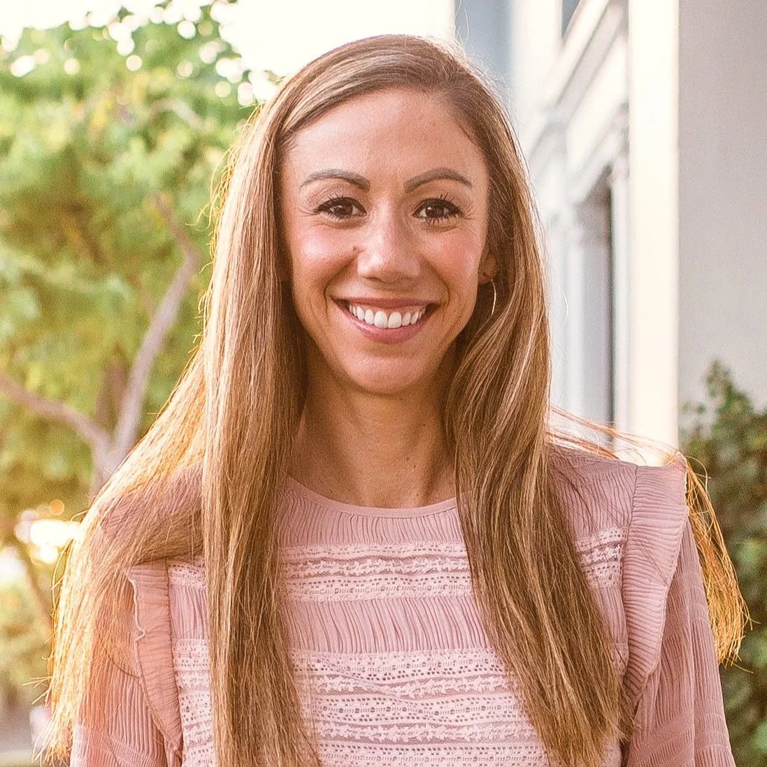 A woman with long brown hair, smiling, standing outdoors with trees in the background, wearing a pink lace dress.