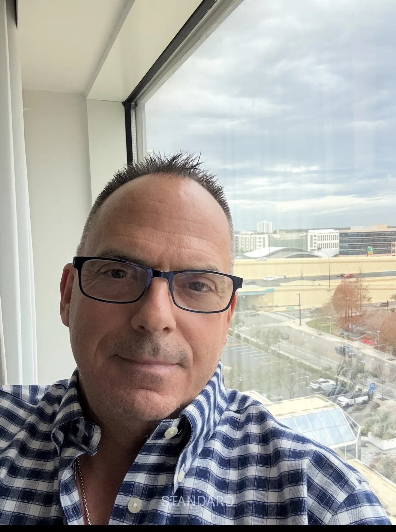 A man with short dark hair, glasses, and a blue plaid shirt taking a selfie in front of a large window with a cityscape in the background. The sky is cloudy, and there are buildings, trees, and a parking lot visible outside.