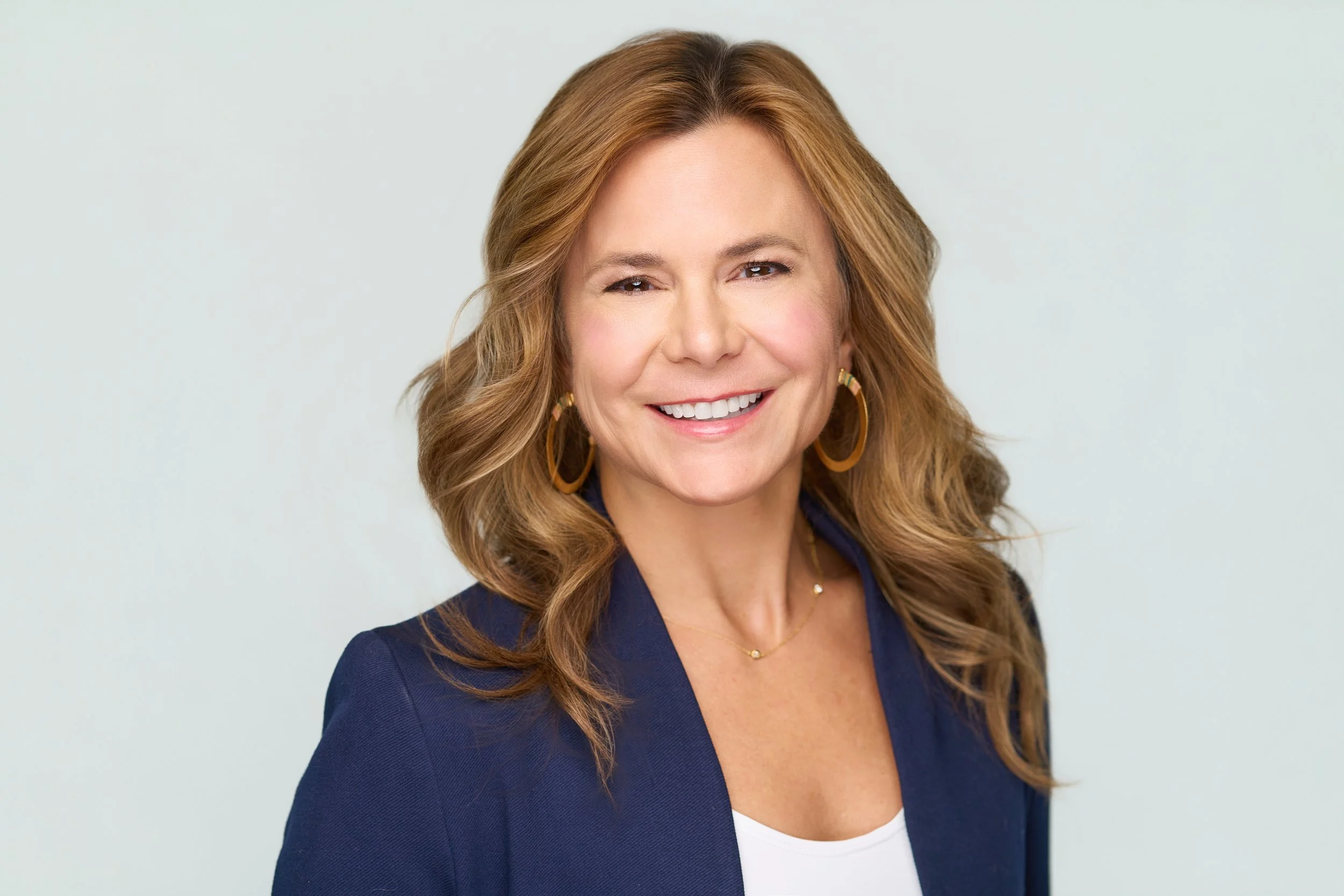 A smiling woman with wavy, shoulder-length light brown hair, wearing gold hoop earrings, a navy blazer, and a white top, standing against a plain light background.