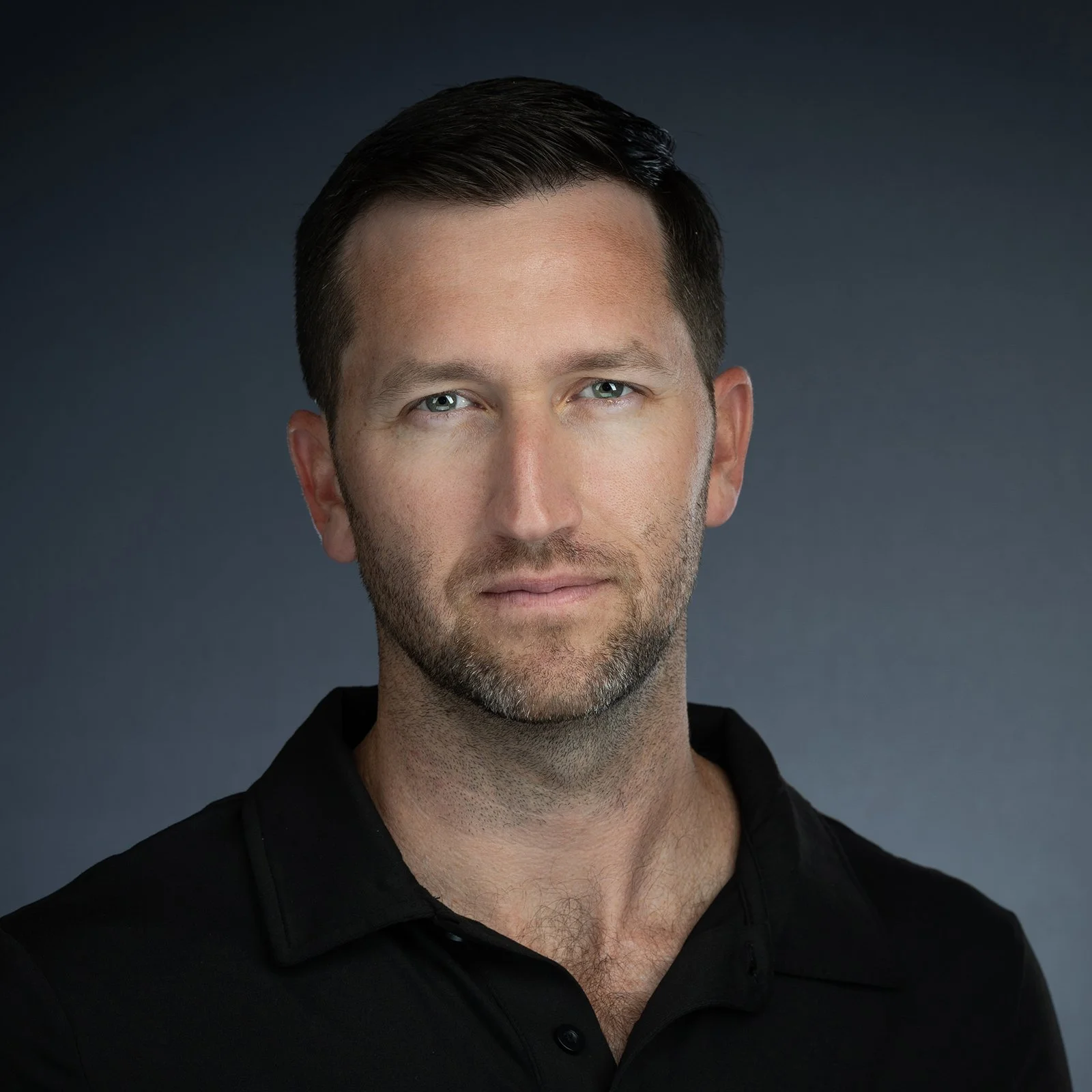 Close-up portrait of a man with dark hair, light eyes, and light beard, wearing a black collared shirt against a dark gray background.