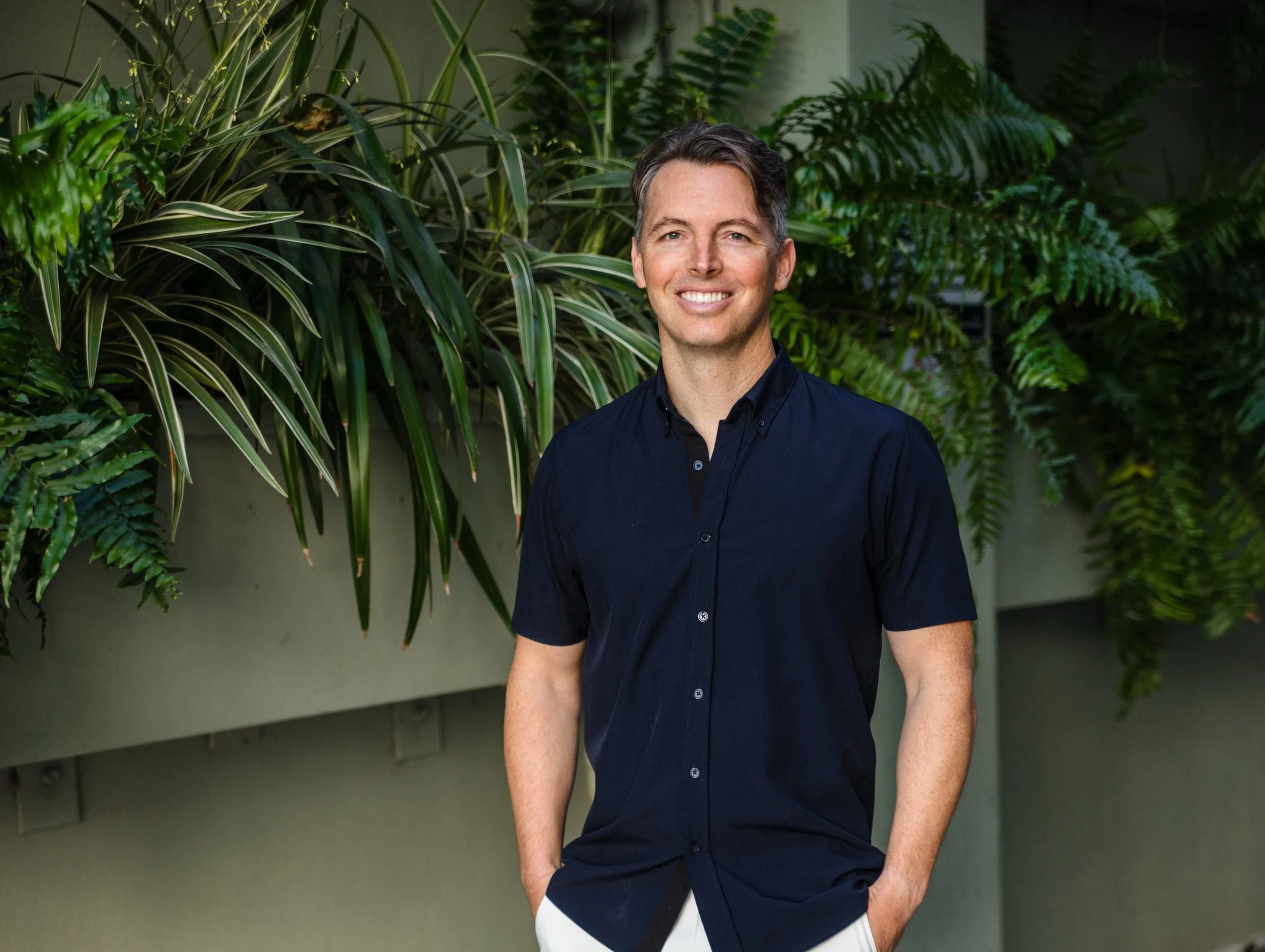 A smiling man with dark hair wearing a navy blue short-sleeve shirt and white pants, standing outdoors in front of green foliage.