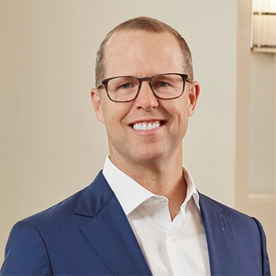 A man wearing glasses and a navy blue suit smiling in an indoor setting.
