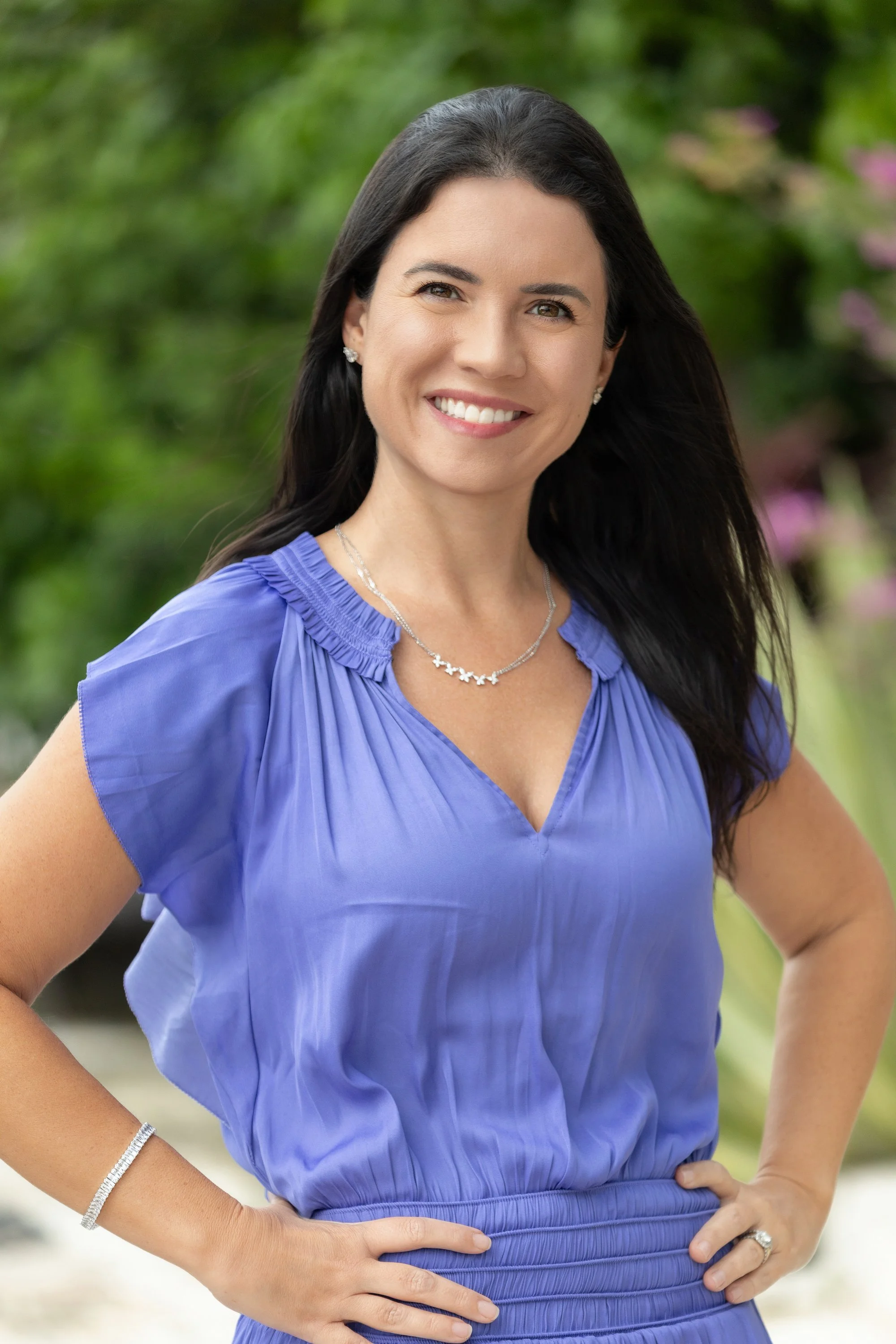 A woman with long dark hair, wearing a blue dress and pearl jewelry, smiling outdoors with green foliage in the background.