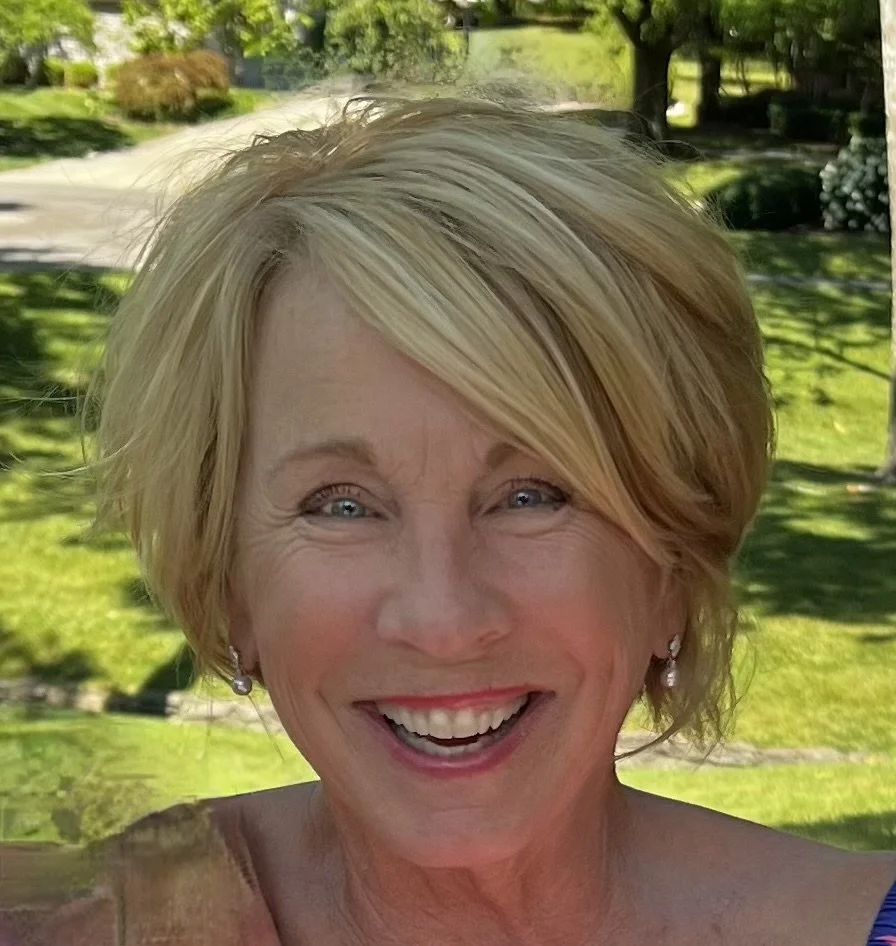 A smiling elderly woman with short blonde hair outdoors in a sunny park.