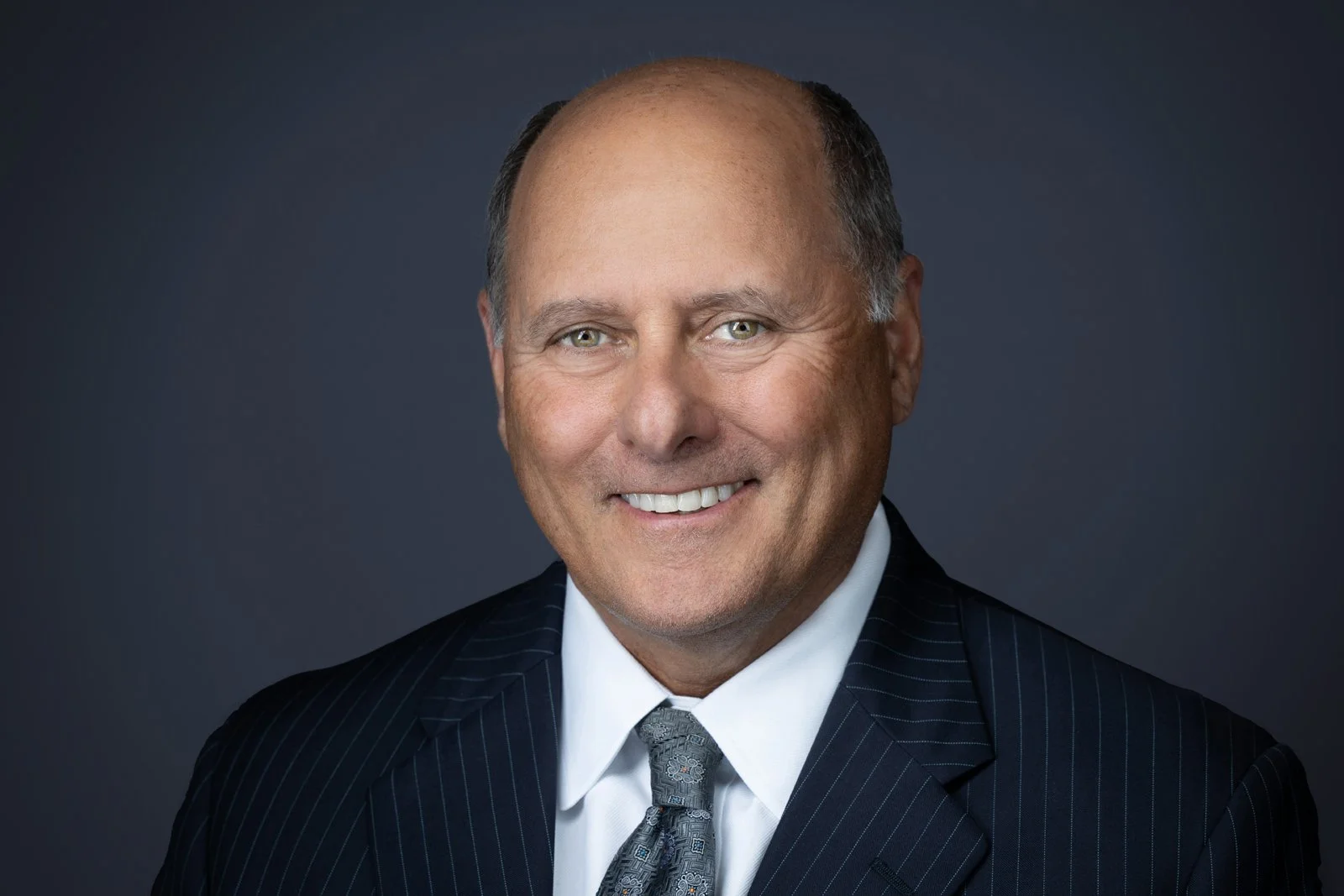 A smiling middle-aged man wearing a dark suit, white shirt, and patterned tie, against a dark grey background.
