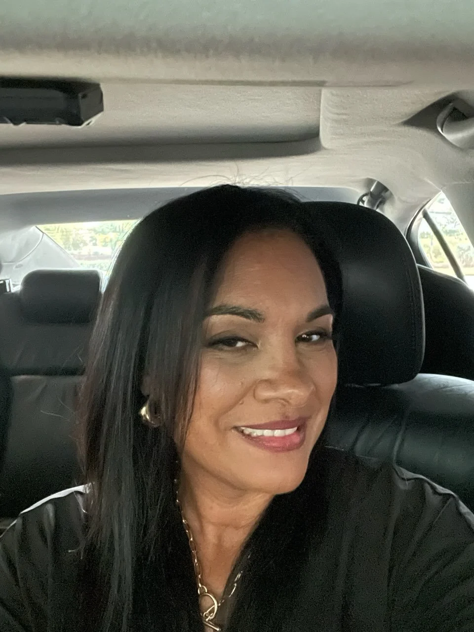 Smiling woman with long dark hair taking a selfie in the backseat of a car