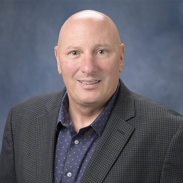 Headshot of a bald man wearing a dark patterned suit and a navy button-up shirt against a blue background.