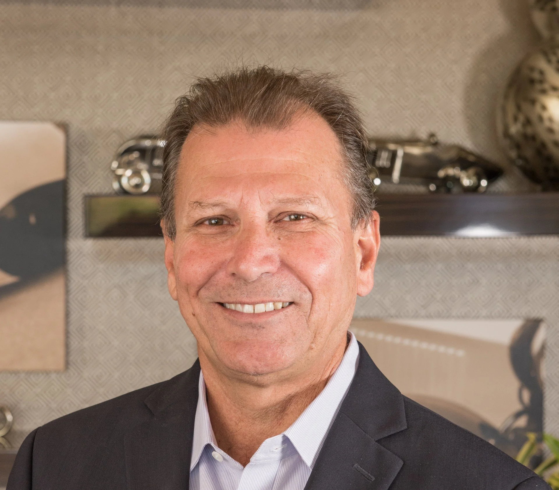 A middle-aged man with short brown hair, wearing a black suit and white shirt, smiling in an indoor setting with shelves and decorative items in the background.