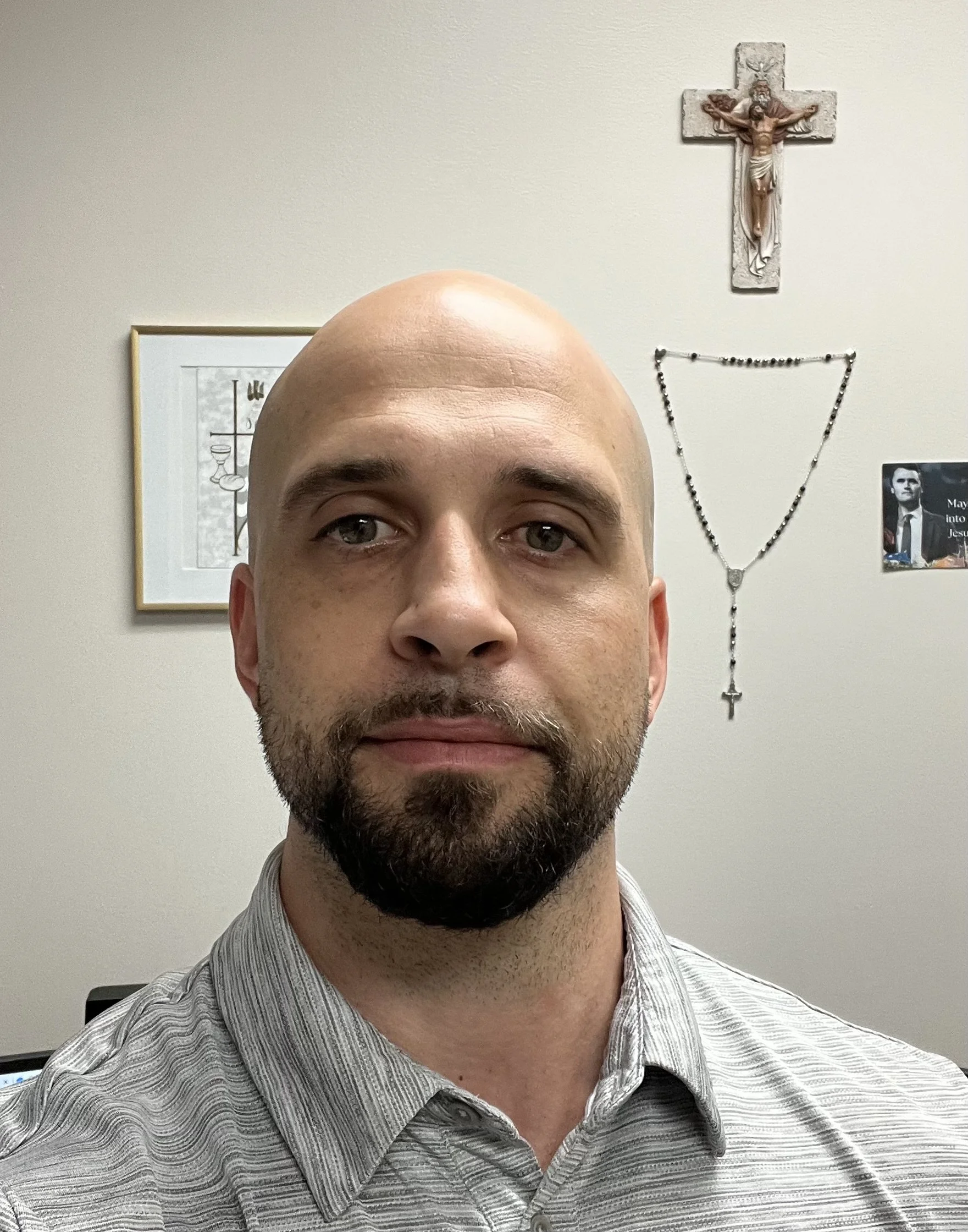 A man with a beard and bald head taking a selfie in front of a religious wall with a crucifix, rosary, and framed picture.