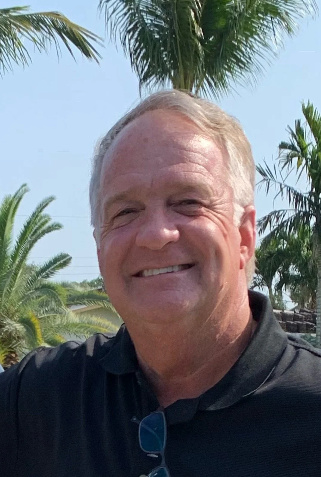 A smiling middle-aged man with short blond hair, wearing a black shirt, outdoors in front of palm trees and clear sky.