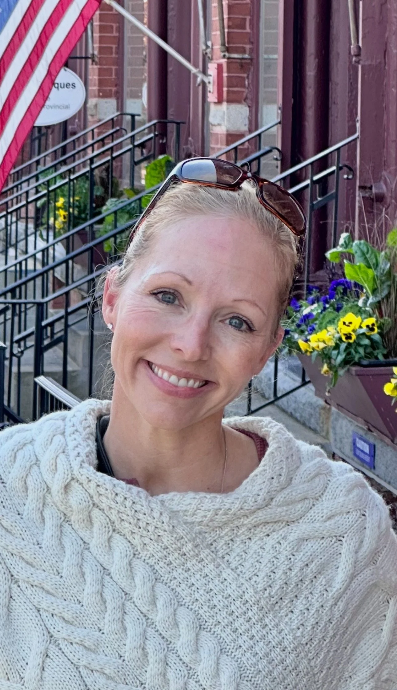 A woman smiling outdoors with sunglasses on her head, wearing a cream-colored knit sweater, standing in front of a building with a flower box filled with purple and yellow flowers.