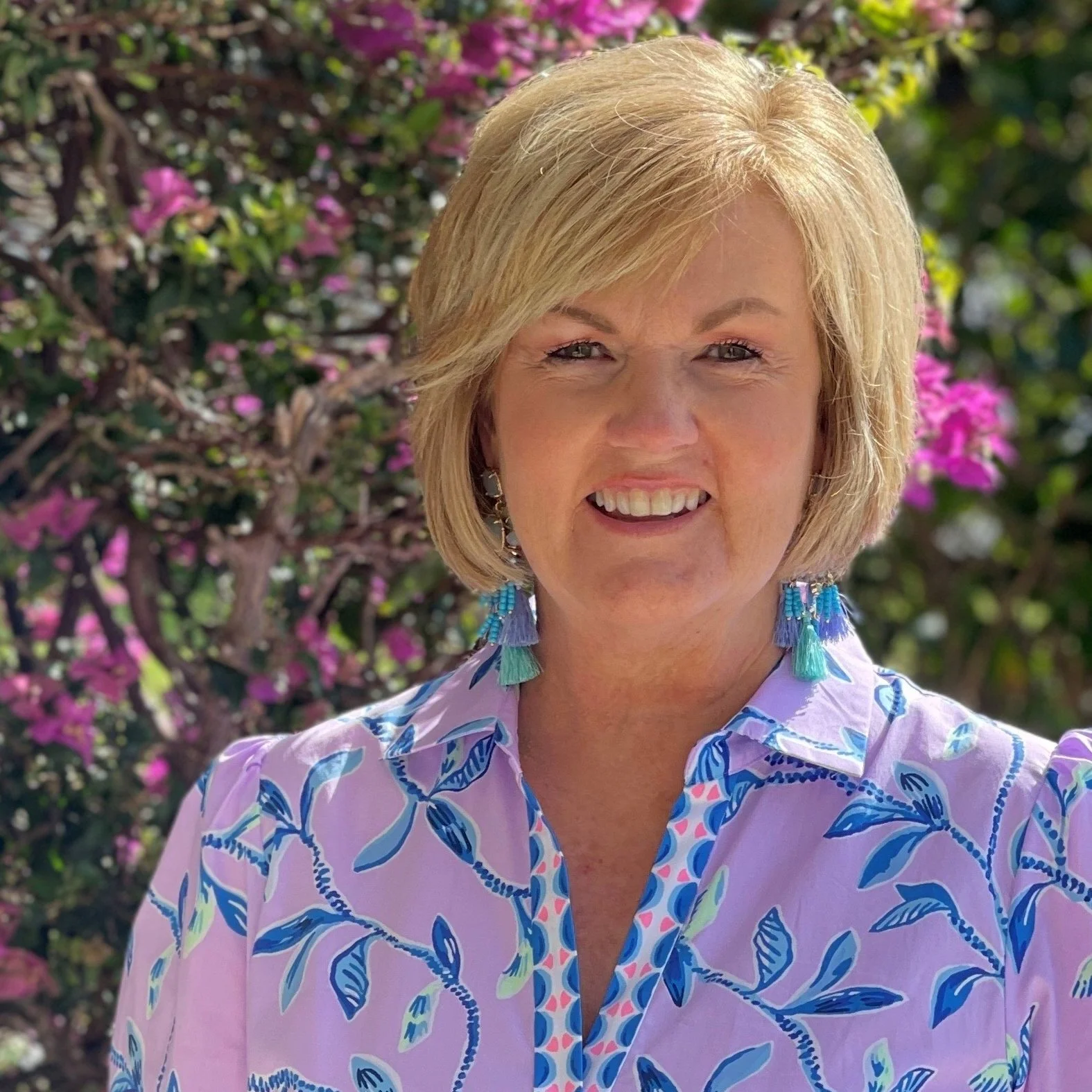 A smiling middle-aged woman with short blonde hair, wearing a pink and blue floral shirt and blue tassel earrings, standing outdoors with pink flowers and green foliage in the background.