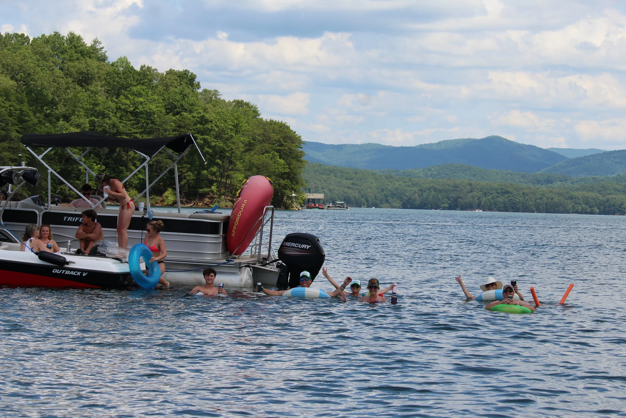 People swimming and relaxing near a boat on a lake with green trees and mountains in the background.